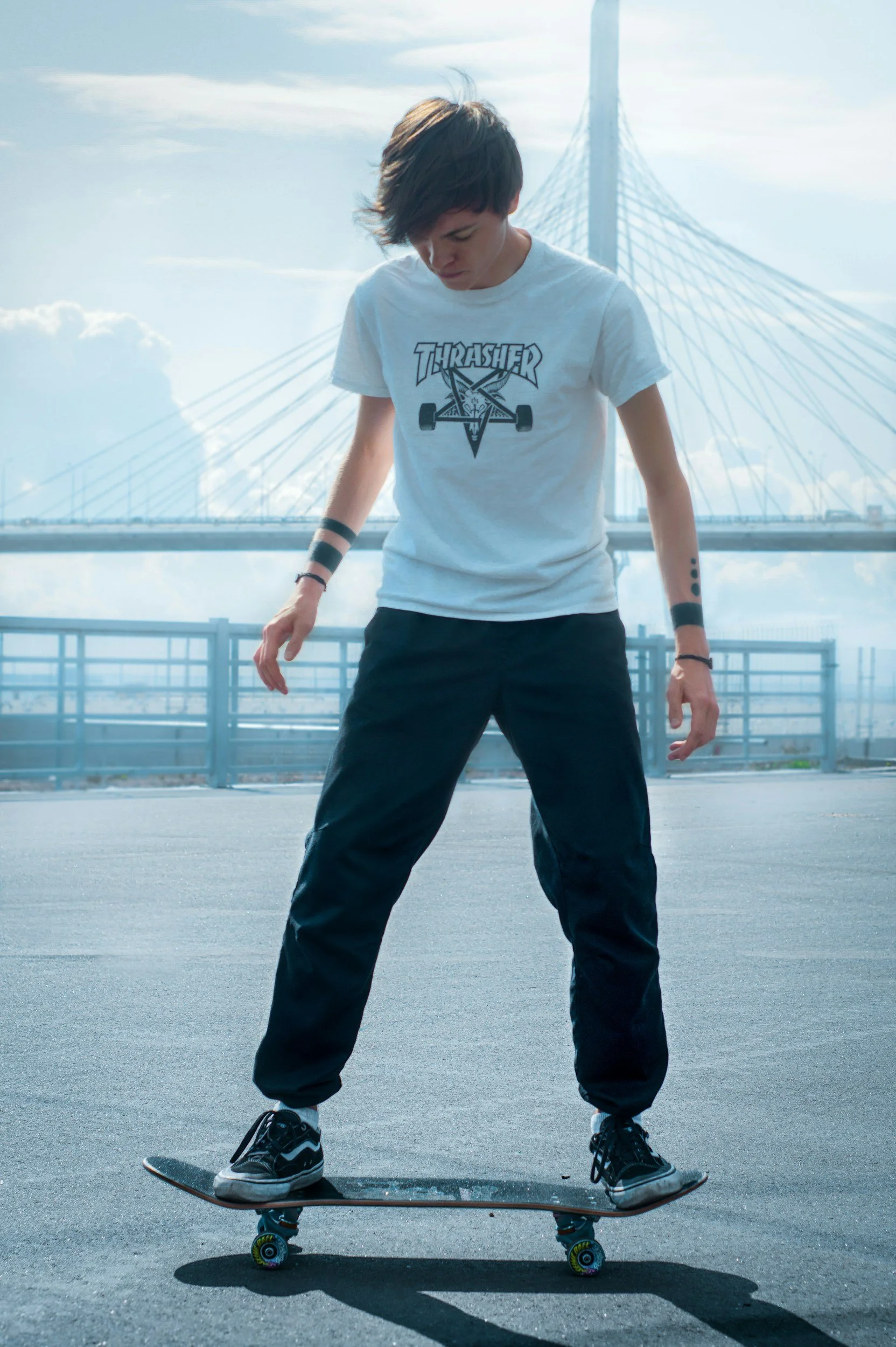 Person skateboarding on a paved surface outdoors, wearing a white T-shirt with 'Thrasher' logo, black pants, and sneakers, with a bridge and blue sky in the background.