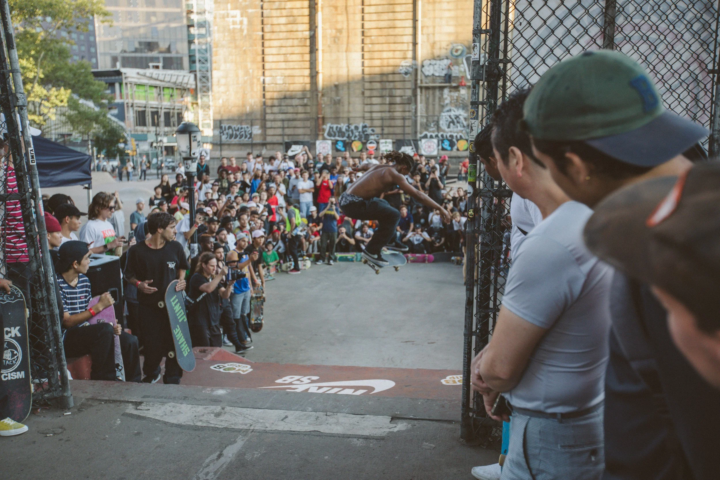 Skateboarder performing a trick over a fence at an outdoor skateboarding event, with a large crowd of spectators and participants watching.