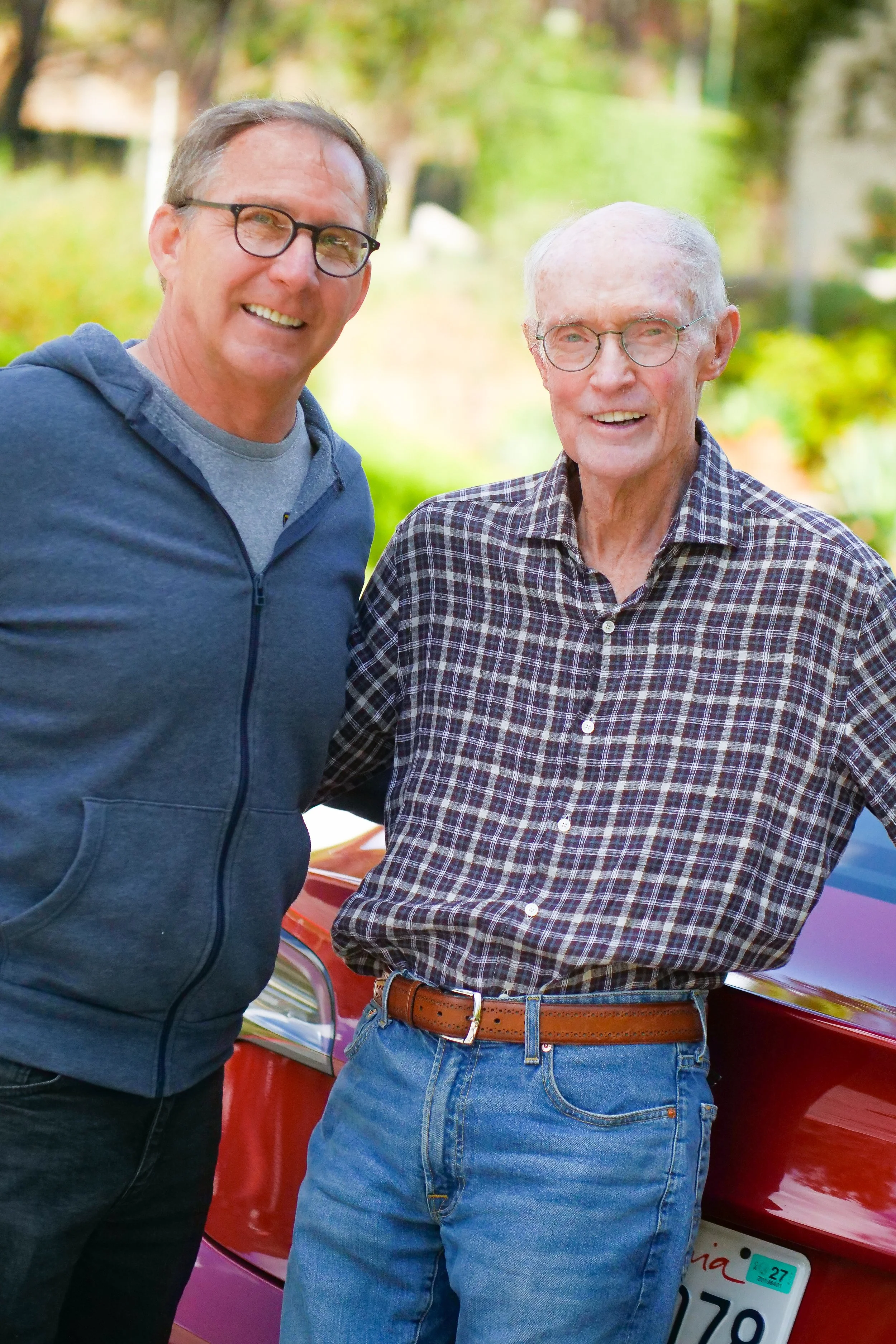 Judge Robert Draper and Pacific Palisades HOA President  stand outdoors in front of a red car, smiling at the camera. The man on the left wears glasses and a dark blue hoodie, and the man on the right wears glasses, a checkered shirt, and jeans.