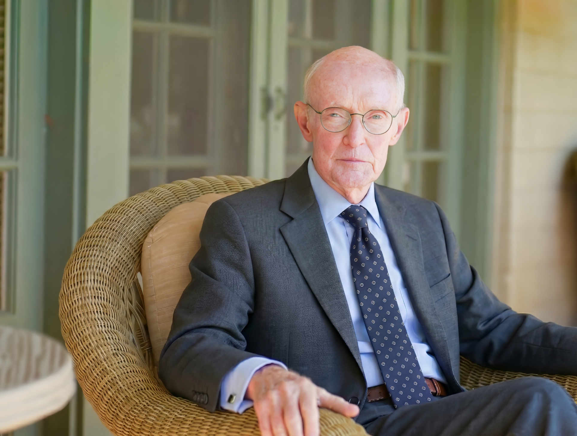 Judge Robert Draper  with glasses wearing a dark suit and a patterned tie, sitting in a wicker chair in front of a porch or veranda, with glass doors and green frame in the background.