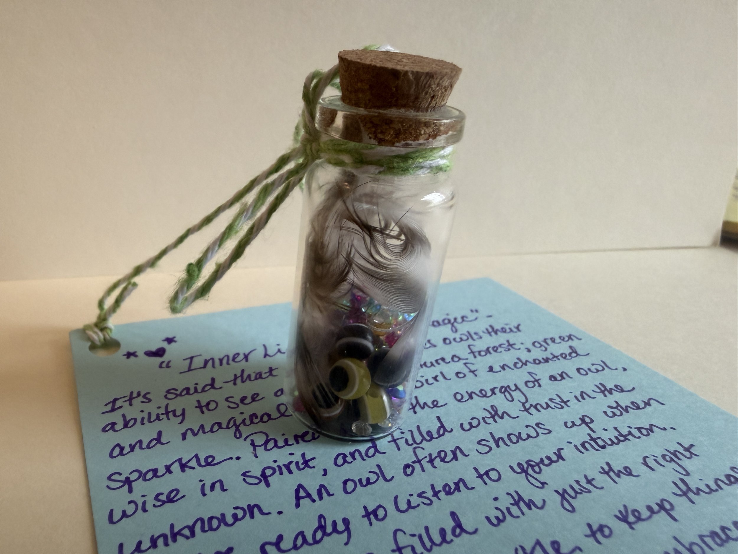 A clear glass jar with a cork lid sitting on a handwritten note. The jar contains black and silver beads, and an eye-shaped pendant is visible at the bottom.