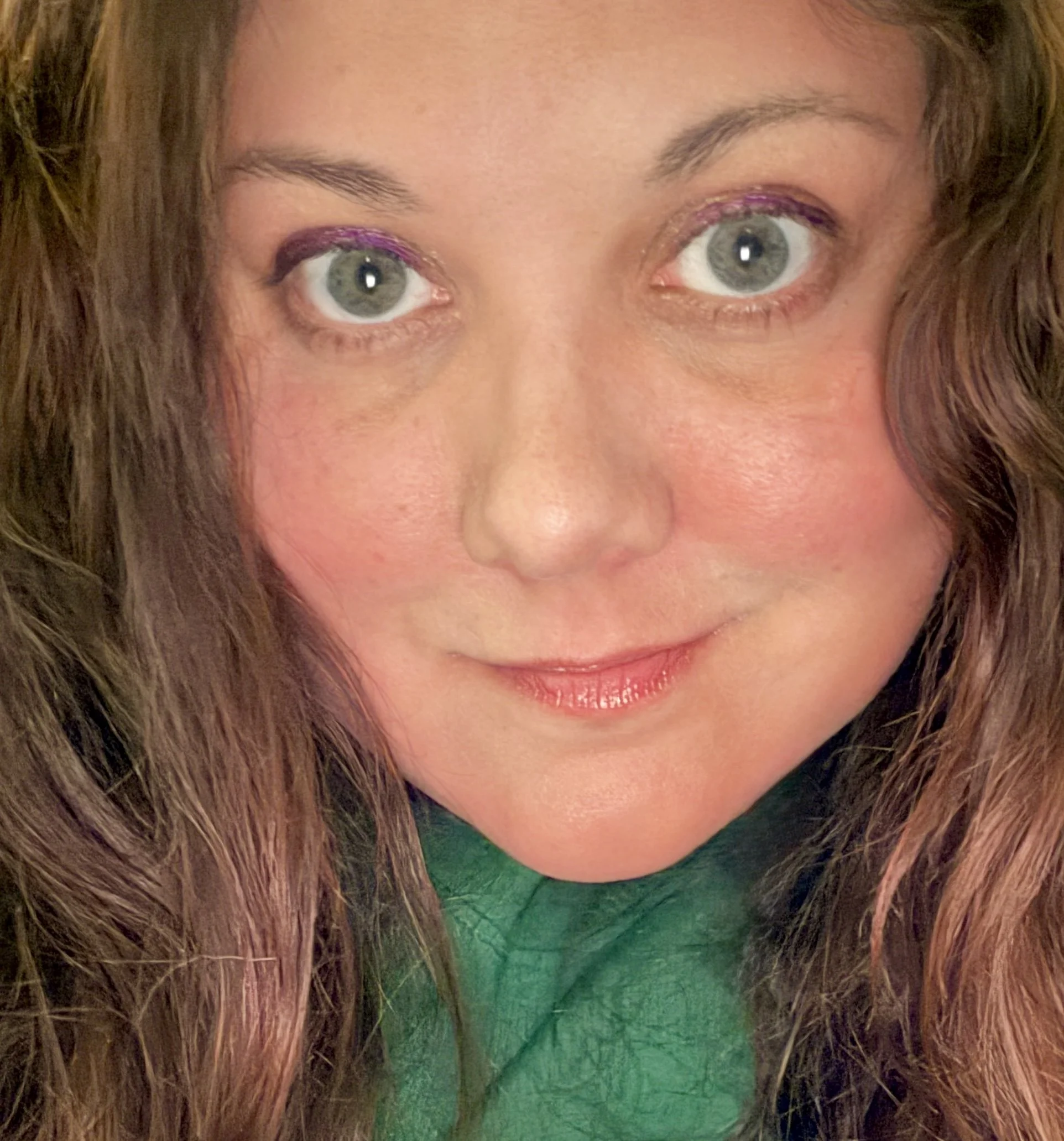 Close-up of a woman with green eyes, long brown hair, and wearing makeup with purple eyeshadow and pink lipstick, smiling slightly.