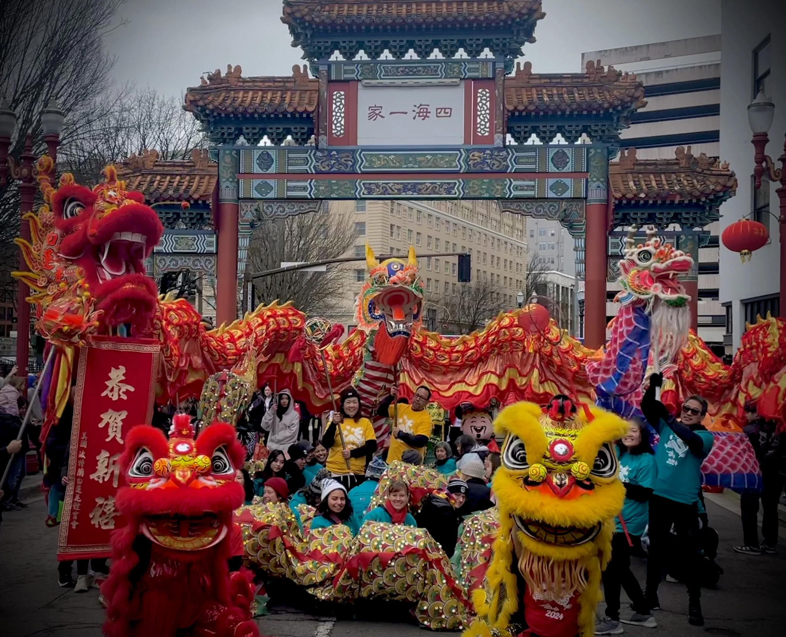A lively outdoor Chinese New Year celebration with people in colorful costumes, traditional lion dance performers, and a decorative Chinese archway with red lanterns and festive banners.