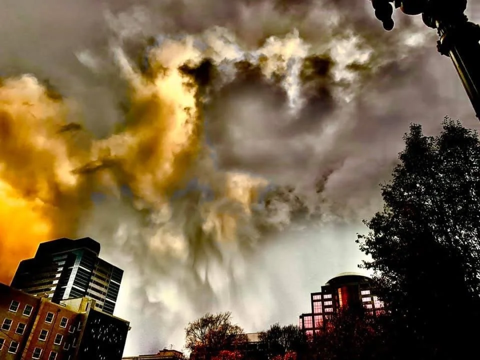 A city skyline with tall buildings and trees under a dramatic, cloudy sky with dark and yellowish clouds.