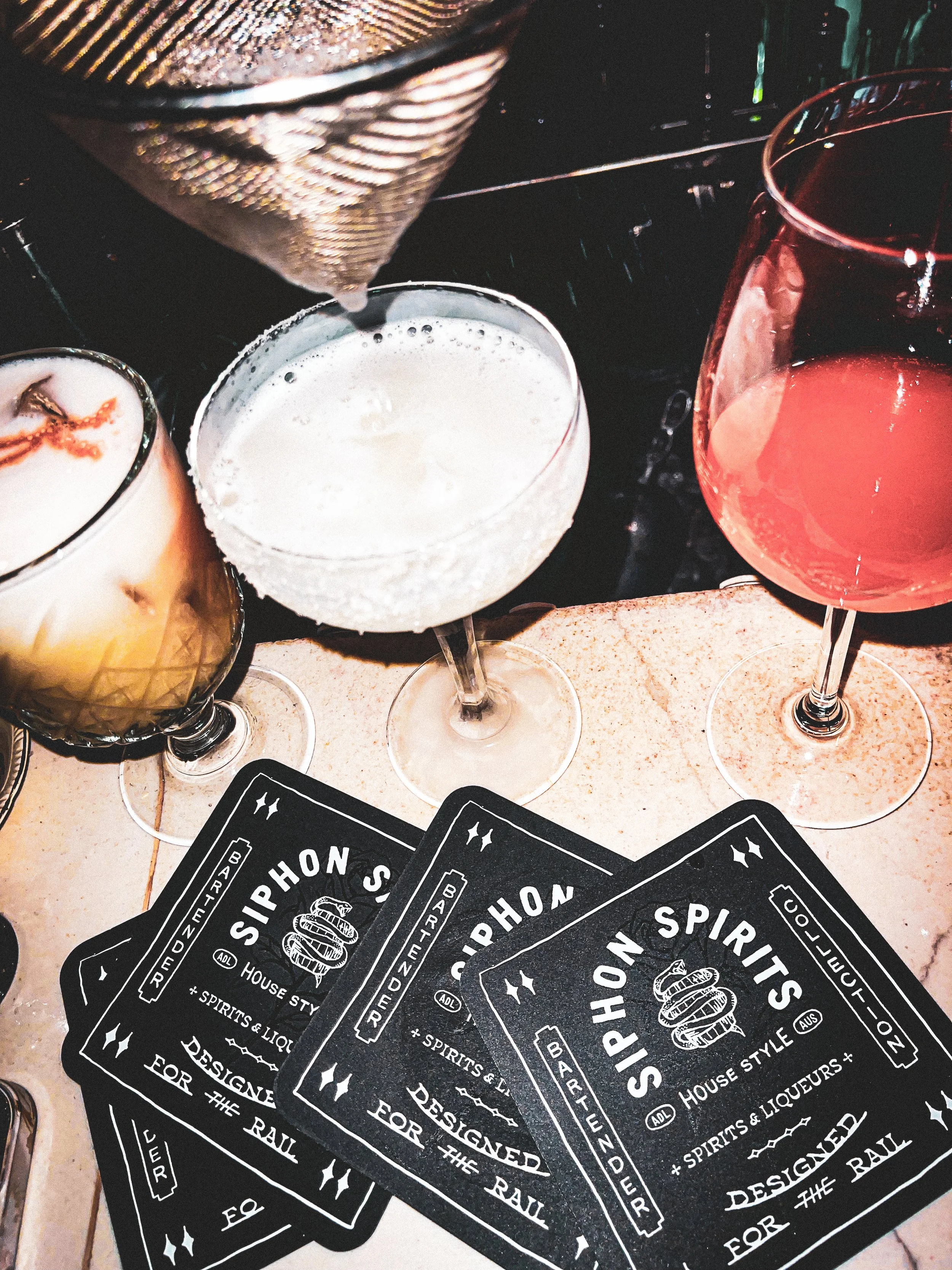 Several black coasters with white text for a bar named Spirit Hons, a menu or promotional material for house spirits and liquors, is laid out on a table. Three cocktail glasses with different drinks are also present; one with a creamy drink garnished with a strip of bacon, a frothy white beverage in a coupe glass, and a red-pink cocktail in a wine glass.