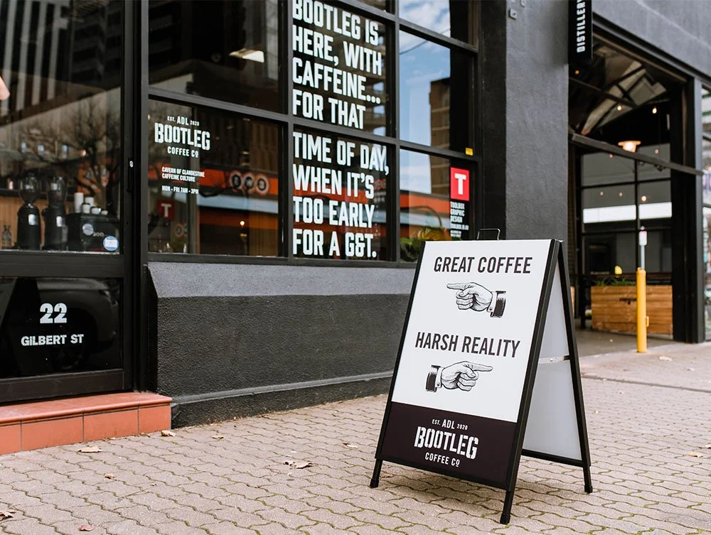 Outdoor sidewalk cafe sign with slogans about coffee, featuring pointing hand graphics and branding for Bootleg Coffee Co, outside a coffee shop.