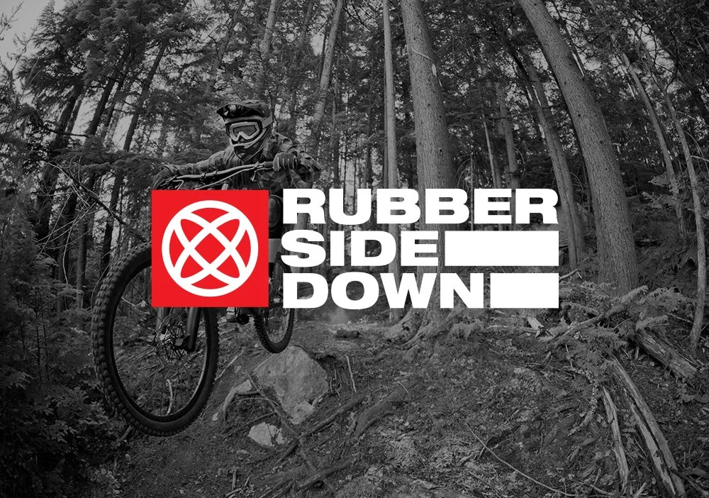 A person mountain biking through a forested trail with tall trees in the background, wearing a helmet and goggles, with the logo and text "Rubber Side Down" superimposed in the foreground.
