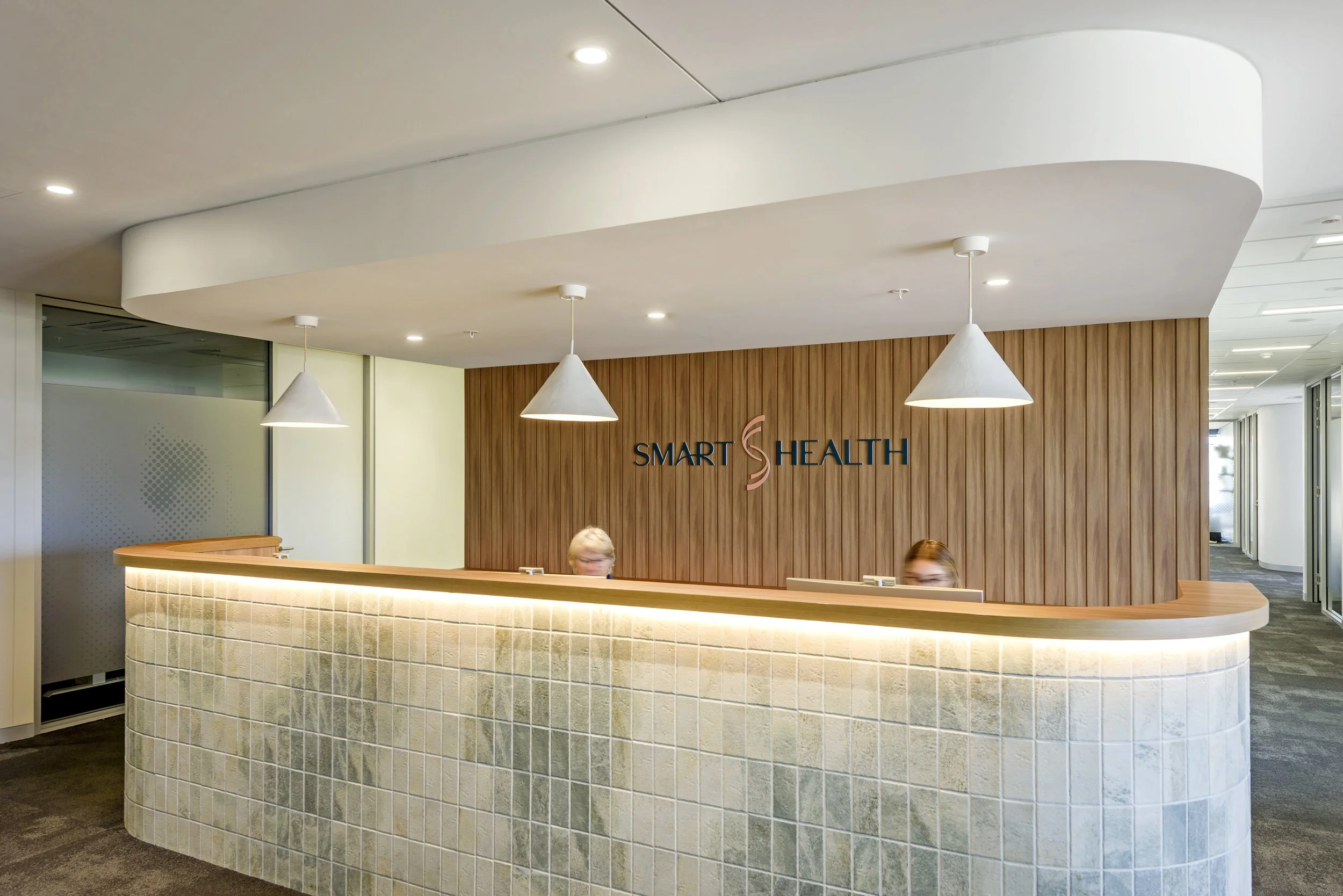 Modern office reception area with a curved desk made of beige tiles, a wooden wall with a 'Smart Health' sign, and two pendant lights hanging from the ceiling.