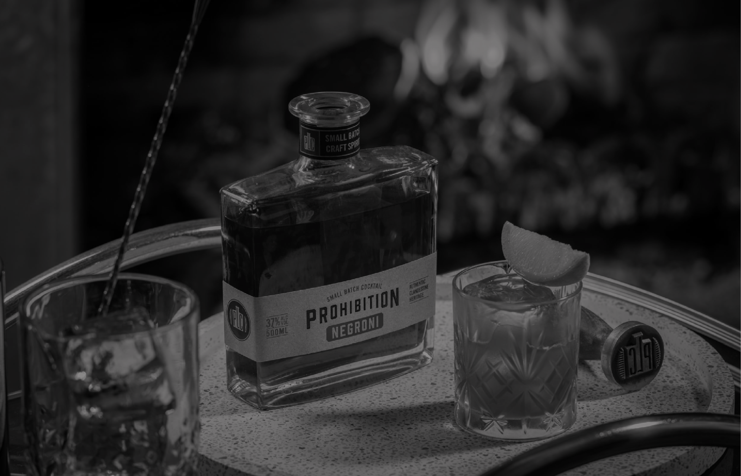 A black and white photograph of a bottle of alcohol labeled "Prohibition" on a table with clear glasses containing drinks and a slice of lemon, some other glasses and objects in the background.