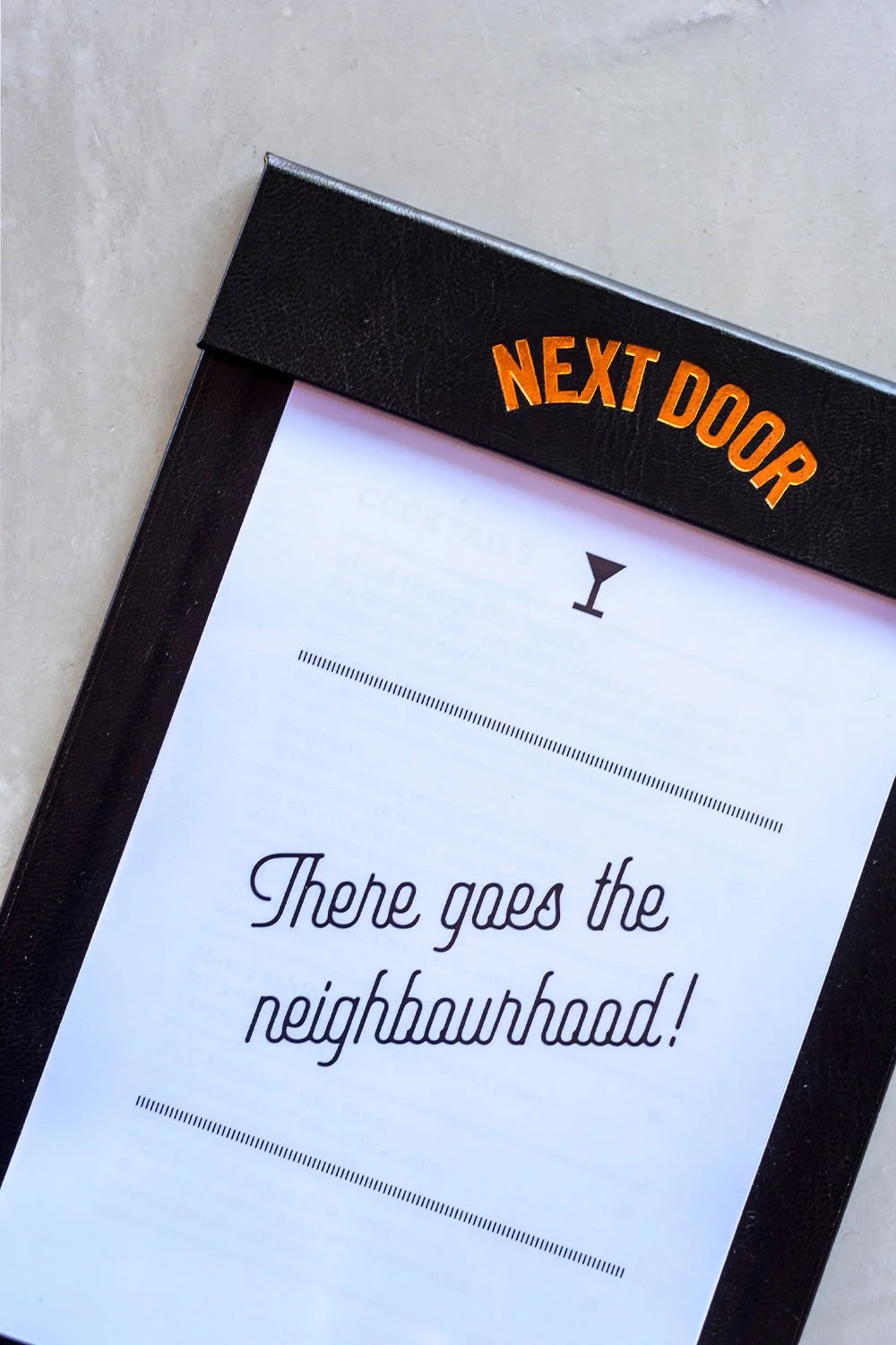 A black menu holder with orange letters reading 'Next Door' on the top, containing a white paper menu with the text 'There goes the neighbourhood!' and a martini glass icon.