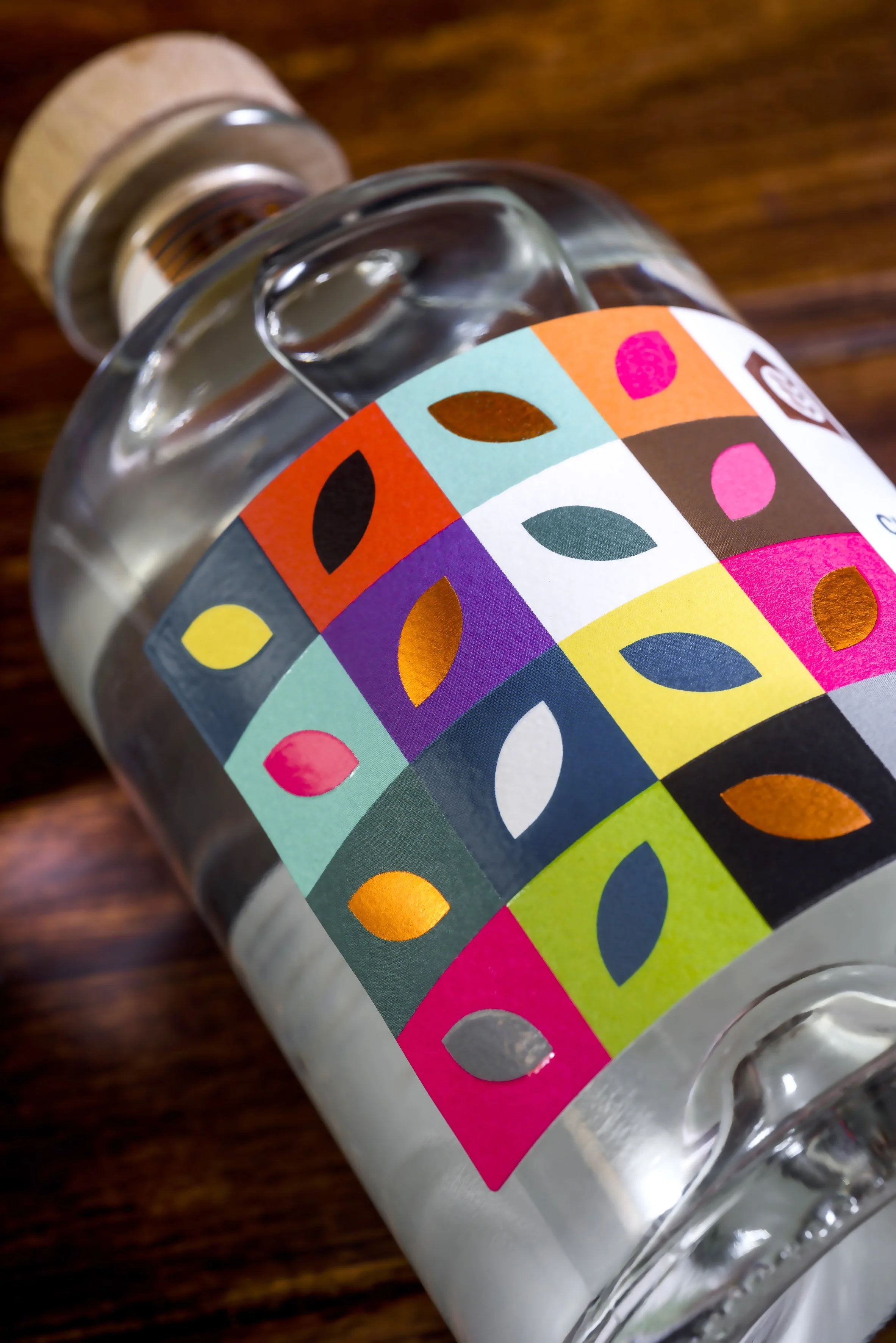 Close-up of a glass bottle with a colorful, patterned label featuring geometric shapes and leaf motifs, lying on a wooden surface.