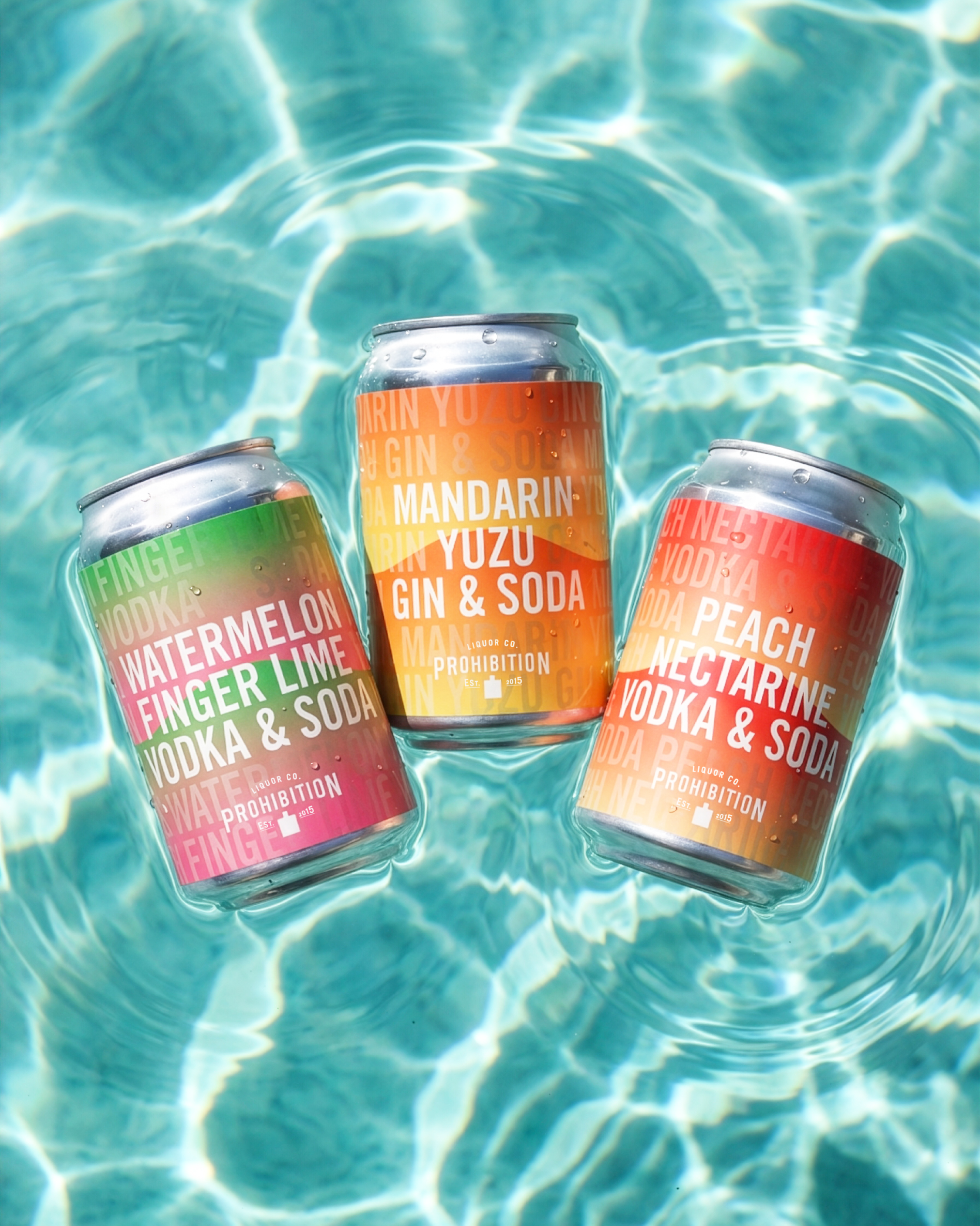 Three cans of flavored soda drinks floating in a swimming pool with clear water and bright sunlight reflections.