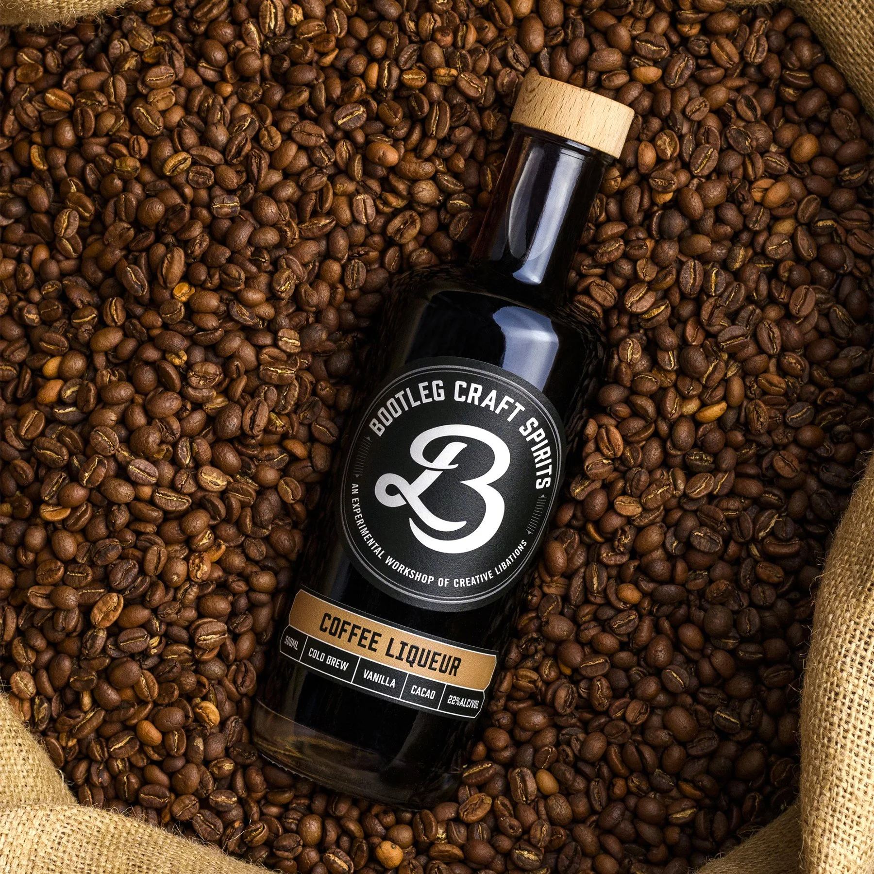 A dark glass bottle of coffee liqueur with a wooden cap, lying on a bed of roasted coffee beans within a burlap-lined container.