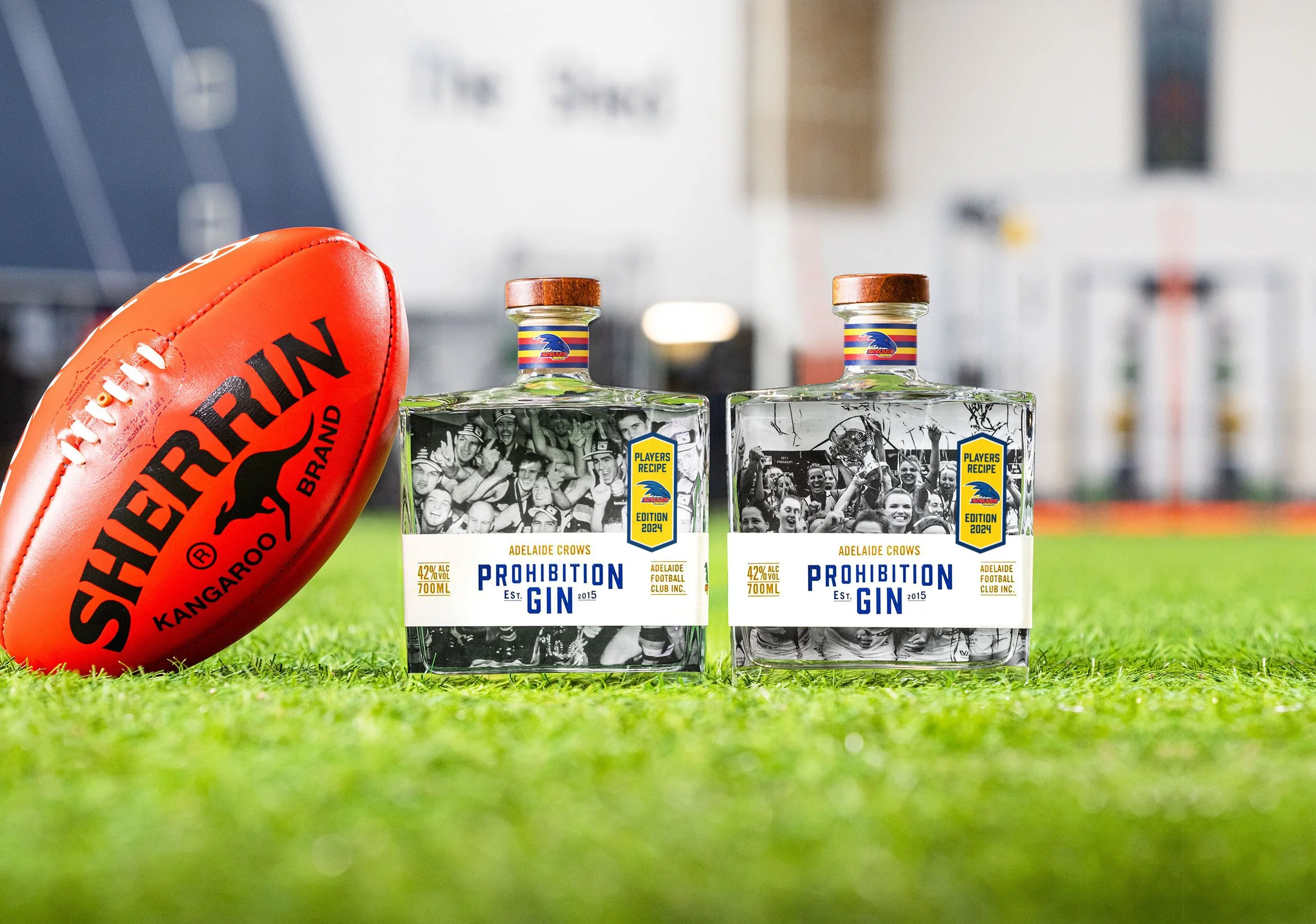 A close-up of an orange football with black text and a kangaroo logo, positioned next to two bottles of Adelaide Crows themed gin on a grassy field with blurred stadium background.