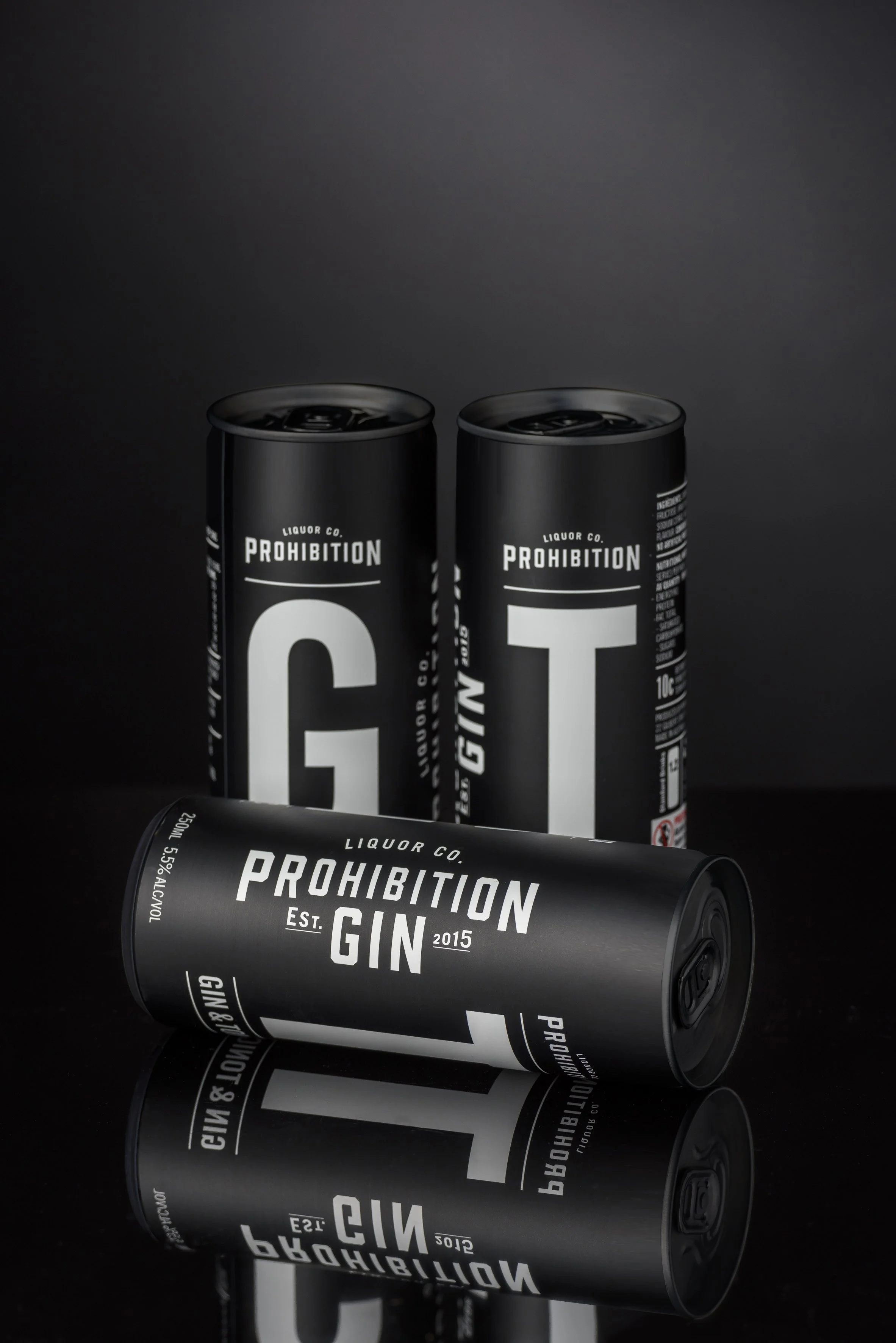 Black cans labeled 'Prohibition Gin' and 'Prohibition' in white text, with a minimalist design, placed on a reflective black surface against a dark background.