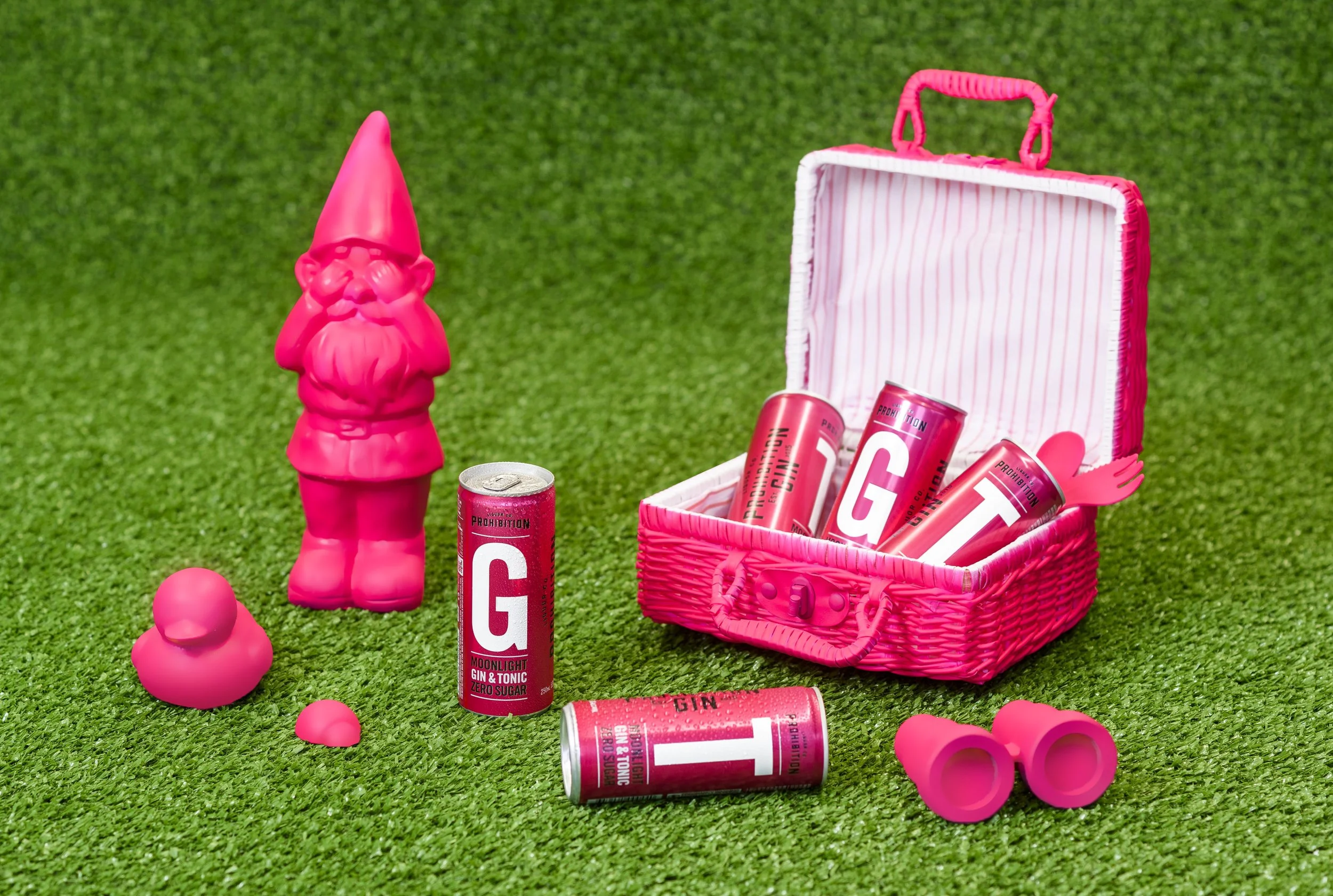 Pink gnome-shaped bottle next to a pink and white striped picnic basket filled with pink cans of gin and tonic, on a green artificial grass surface with scattered pink rubber balls.