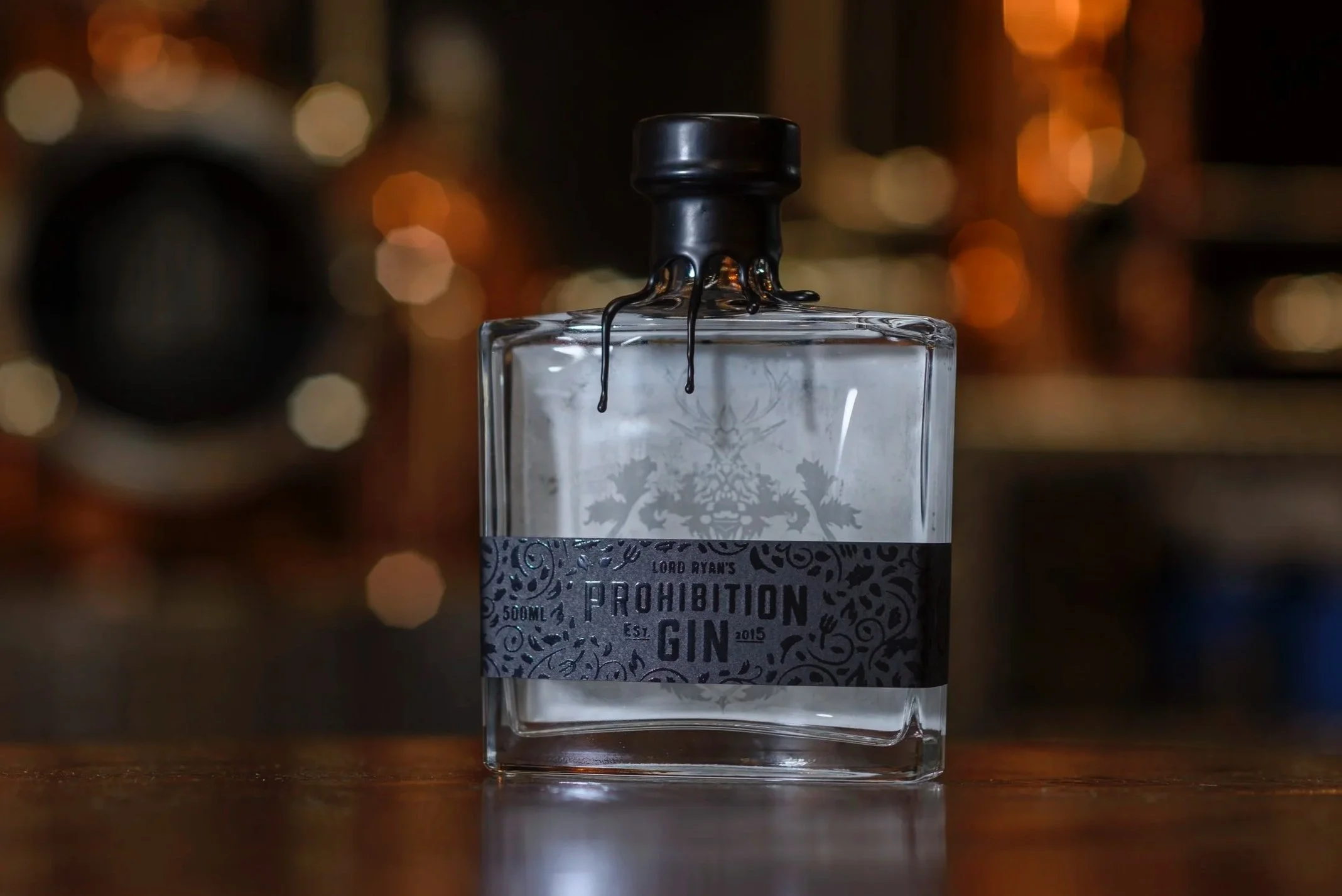 A clear glass bottle labeled 'Prohibition Gin' with black liquid overflowing from the top, against a blurred background of warm-colored lights.
