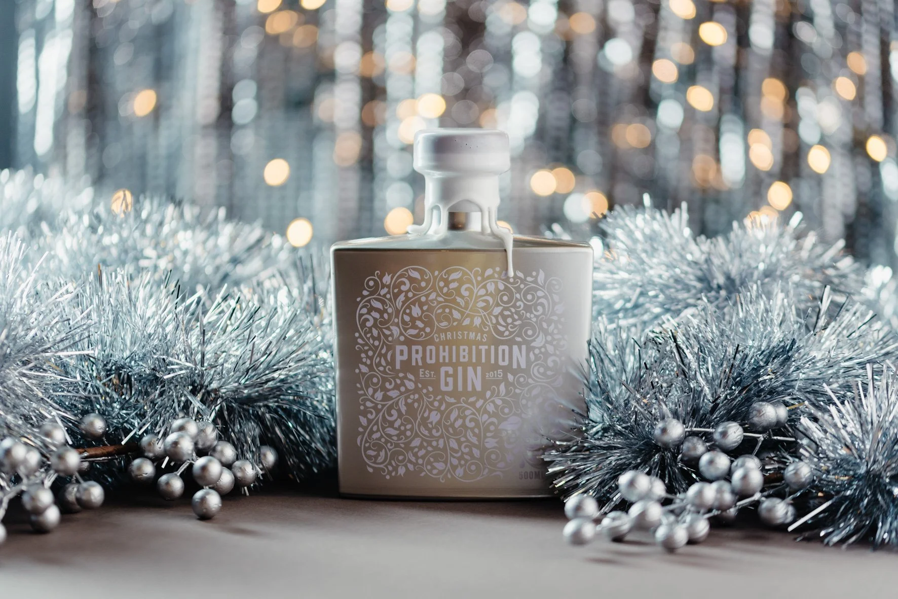 A decorative Christmas-themed display featuring a white bottle labeled 'Christmas Prohibition Gin' surrounded by silver tinsel and silver beads, with a blurred background of festive lights.