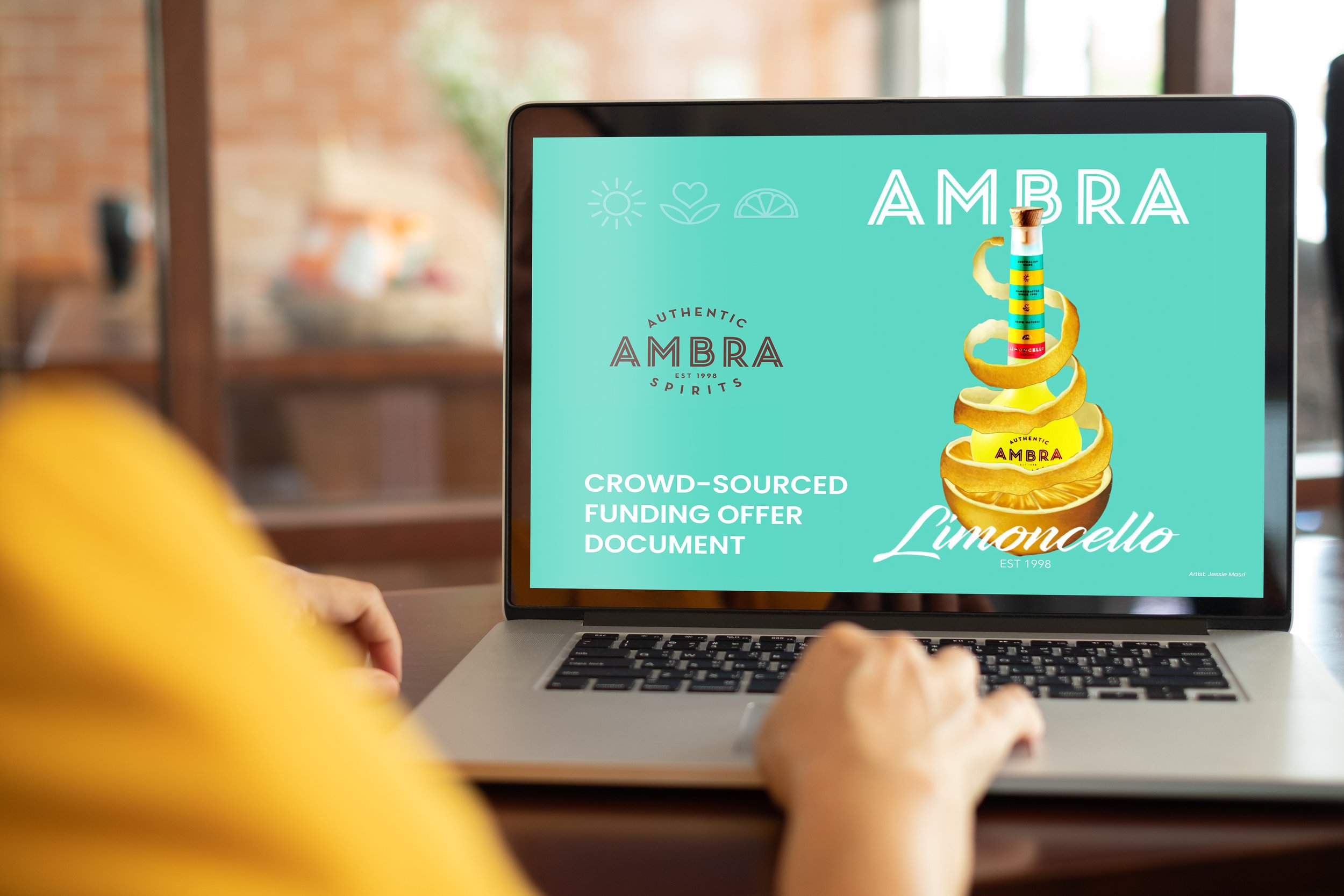 Close-up of a laptop screen displaying a colorful advertisement for Ambra Limoncello, featuring an illustration of a lemon peel spiral and a bottle of Limoncello, with text promoting a crowd-sourced funding offer.