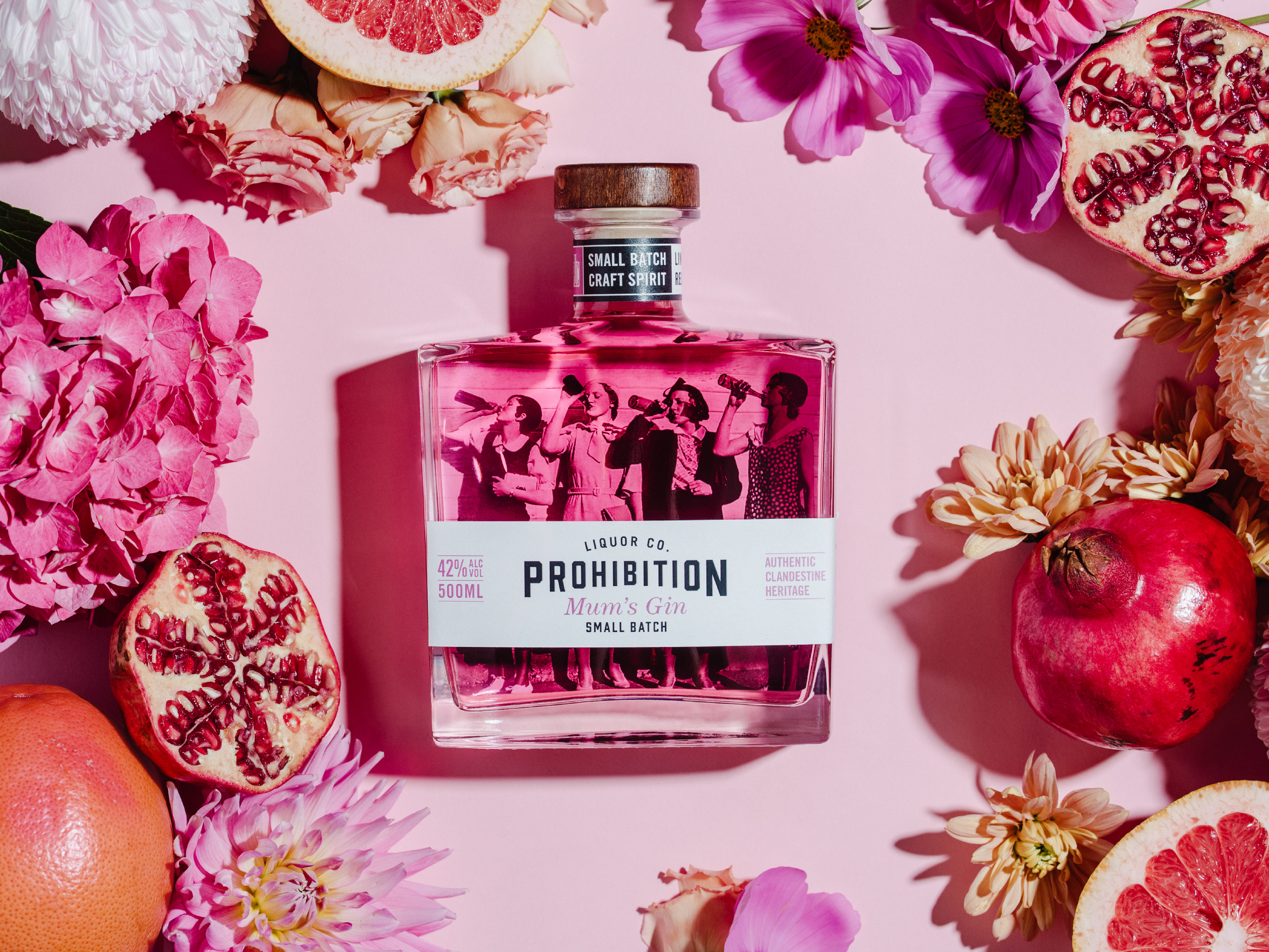 A bottle of pink gin called 'Prohibition' surrounded by pink and red flowers and pomegranates on a pink background.