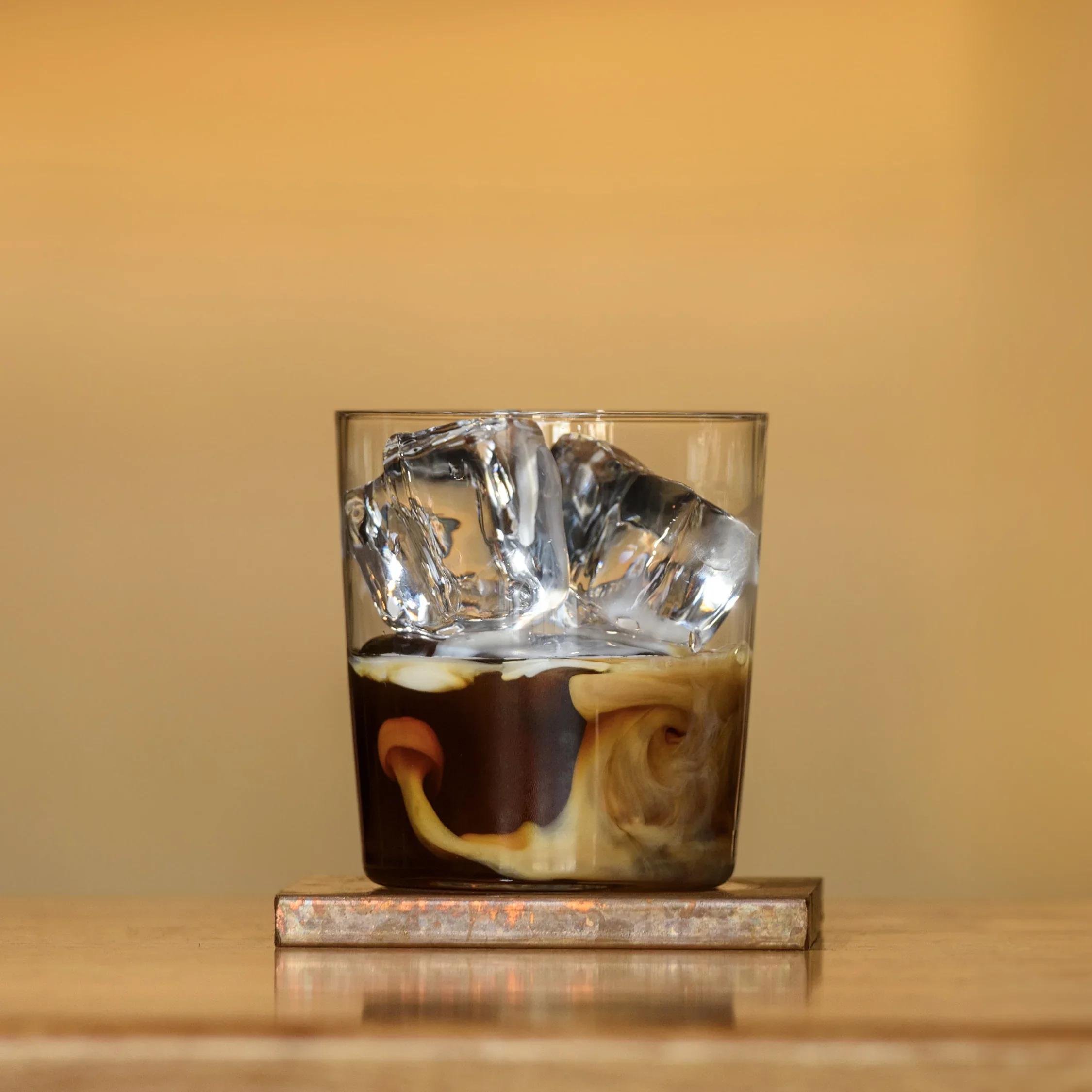 Glass with ice cubes and dark liquid, placed on a marble or stone coaster on a wooden surface.