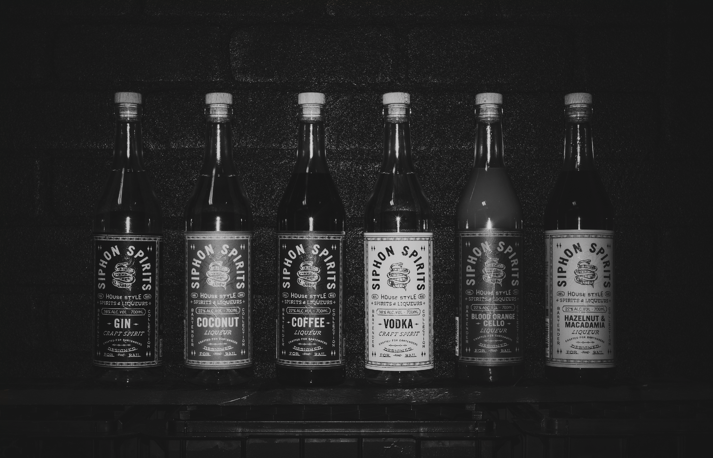 Black and white photo of six liquor bottles with labels including gin, coconut liquor, coffee liquor, vodka, blood orange, and hazelnut macadamia, placed on a dark shelf with a dark background.