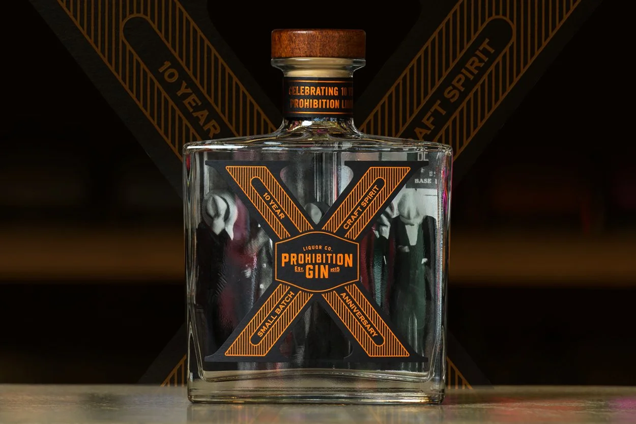 A clear glass bottle of Prohibition Gin with a wooden cap, featuring orange and black labels, and a dark background with diagonal stripes and text celebrating the 10-year anniversary of the prohibition law.
