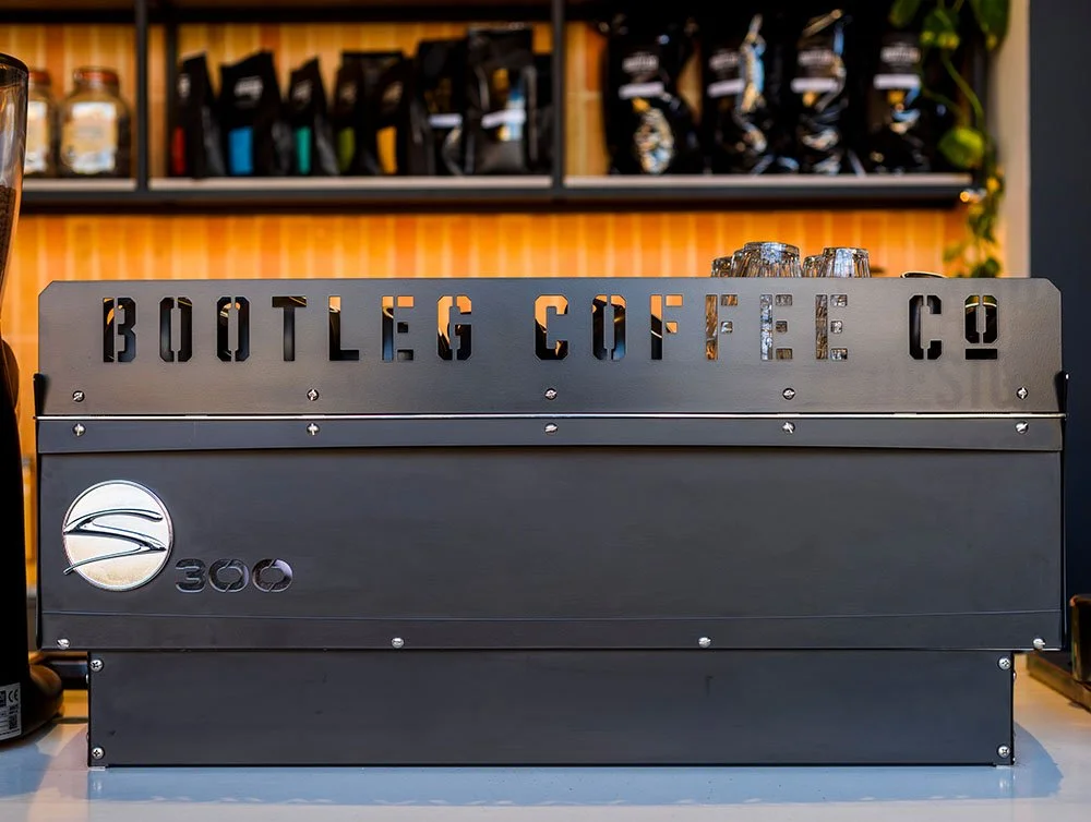 A modern coffee vending machine with the sign 'Bootleg Coffee Co' and a logo with a wave symbol, placed on a counter with cups on top, in a coffee shop setting.