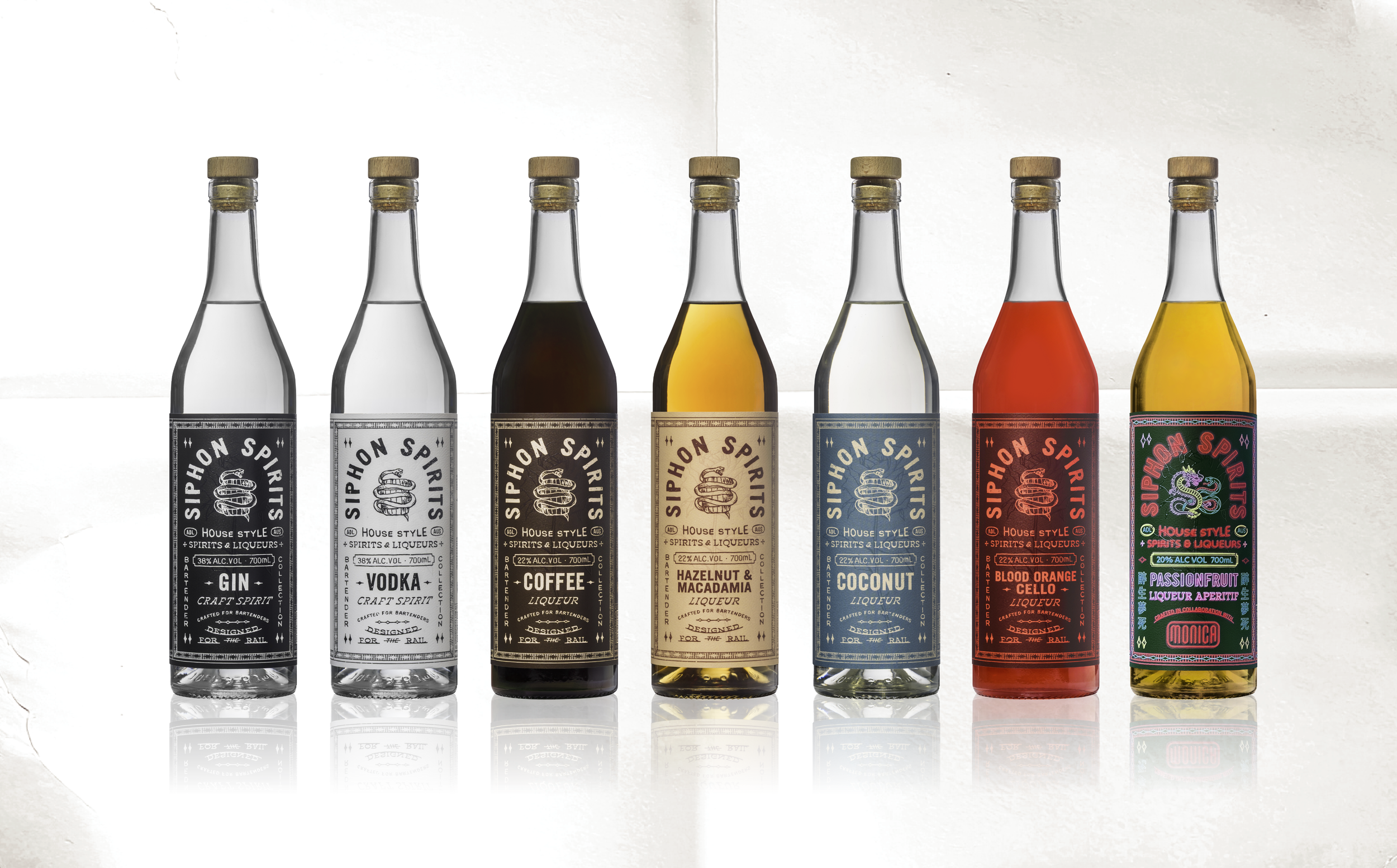 Six bottles of craft spirits with colorful labels, including gin, vodka, coffee liquor, hazelnut macadamia liqueur, coconut liqueur, and blood orange cello, all with cork stoppers, on a reflective surface.