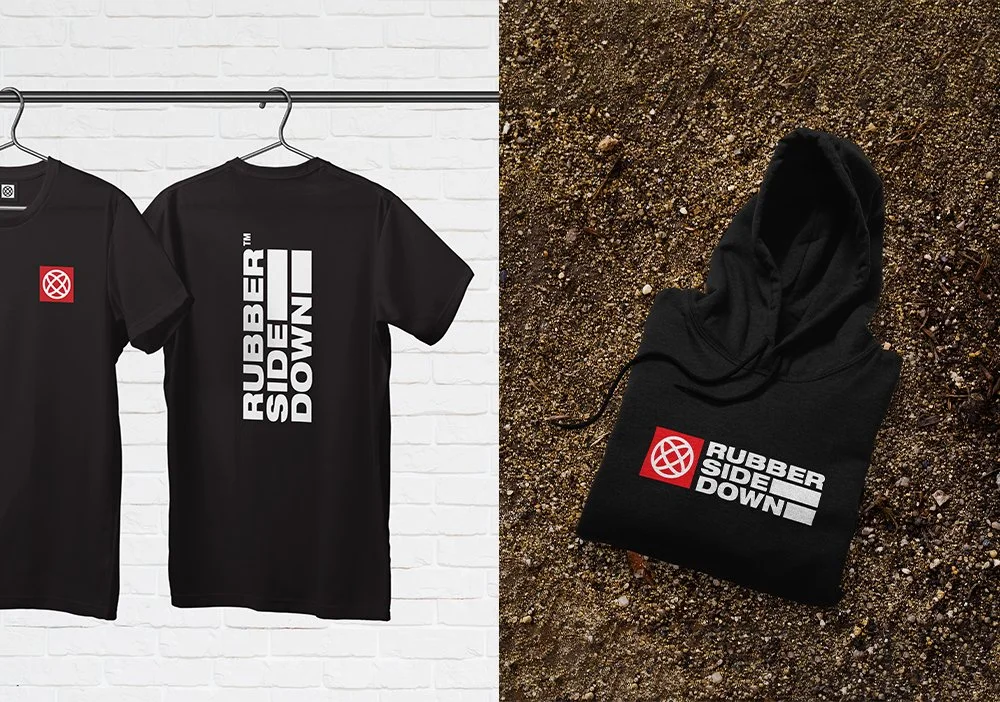 Two black T-shirts hanging on a rack, one with a red logo and the other with white text that reads "RUBBER SIDE DOWN!"; a folded black hoodie lying on dirt ground, with a logo and text that says "RUBBER SIDE DOWN!".