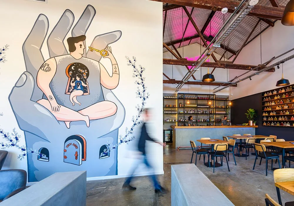 Interior of a modern café with a mural of a person holding a key, sitting in front of a large hand, with a tiny house at the bottom. The café has a bar with shelves full of bottles, tables and chairs, and industrial ceiling lighting.