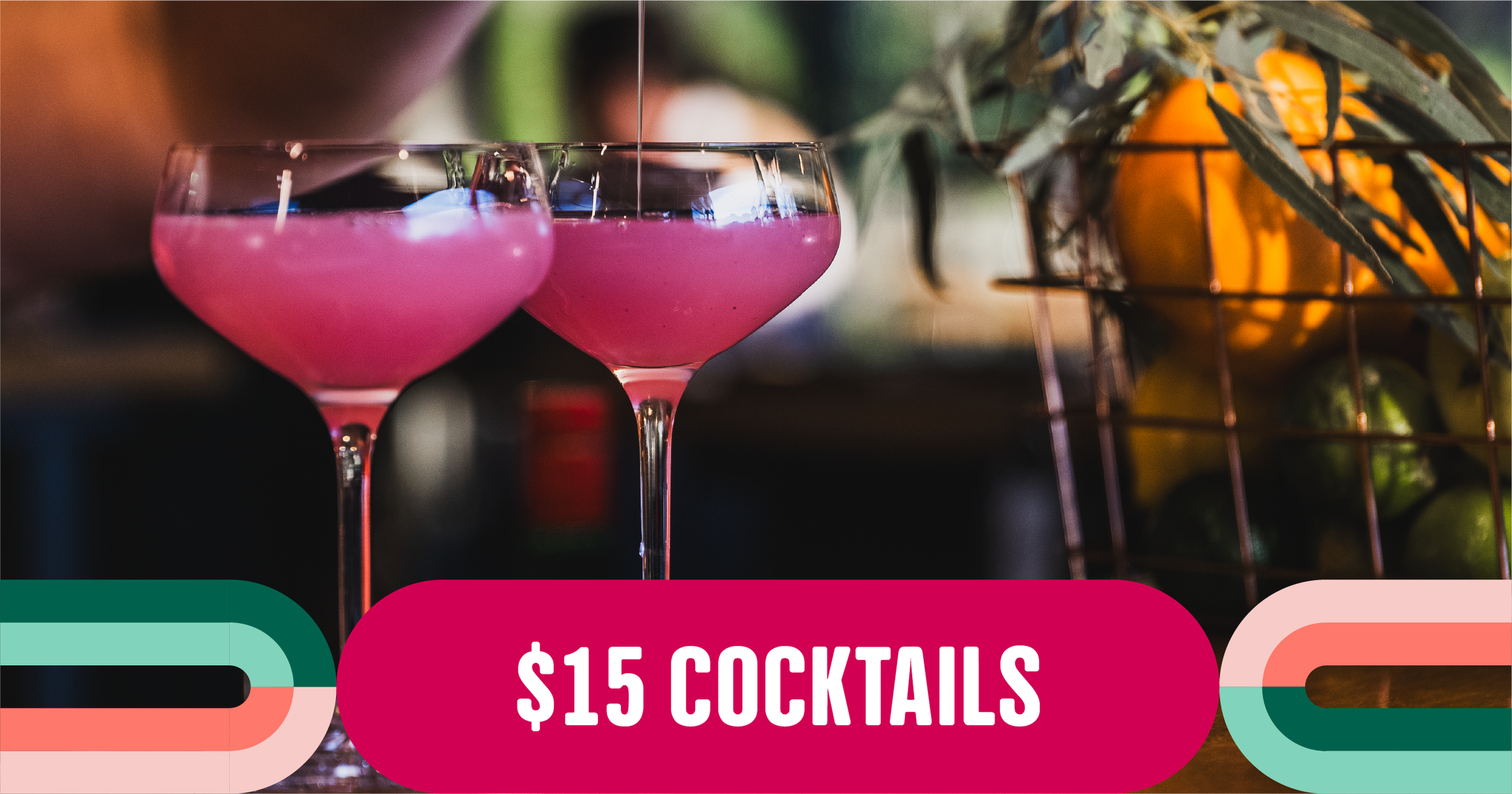 Two pink cocktails in coupe glasses with ice, with a blurred colorful background including a basket of oranges and greenery.