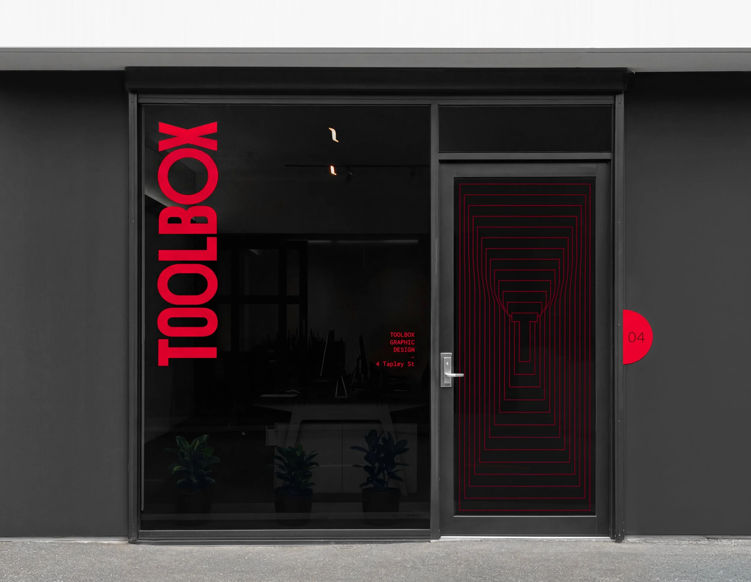 Modern black storefront window with vertical red text reading 'TOOLBOX' and side window featuring a stylized red outline of a toolbox with the number 04.