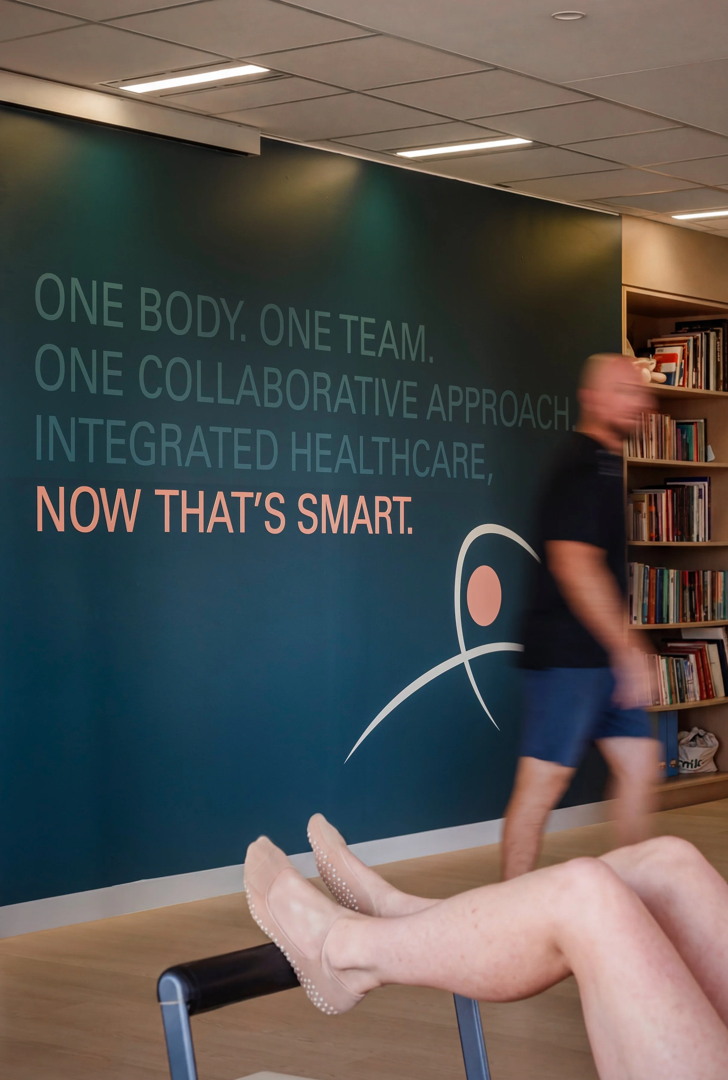 A scene from inside a building, showing a woman engaged in a chiropractic adjustment on a patient’s ankle while sitting on a chair. The background displays a wall with motivational text about teamwork and healthcare, and a bookshelf with various books.