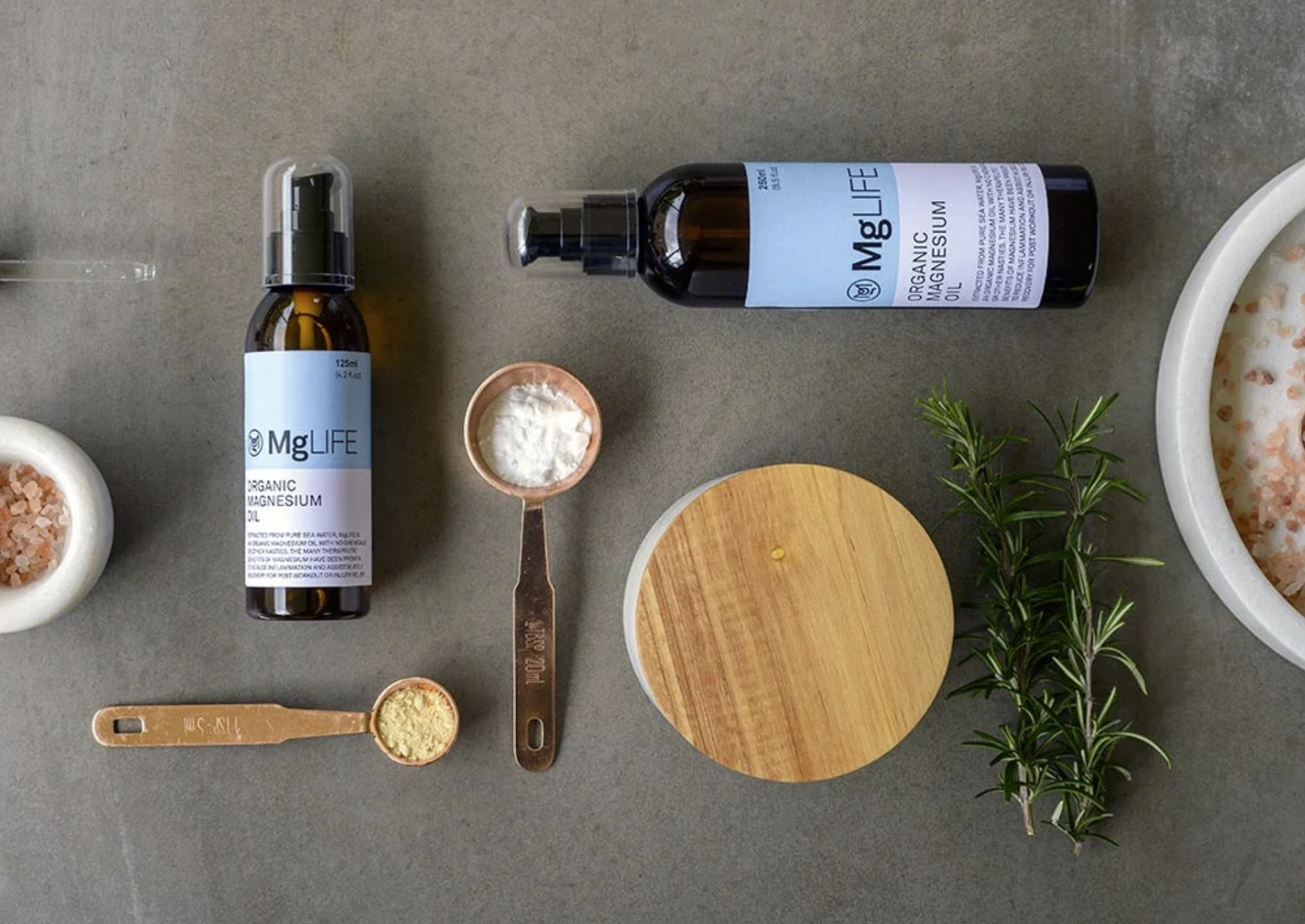 Top-down view of a flat surface with bottles of magnesium oil, small bowls of powder, measuring spoons, a wooden lid, a sprig of greenery, and a large bowl of pink Himalayan salt.