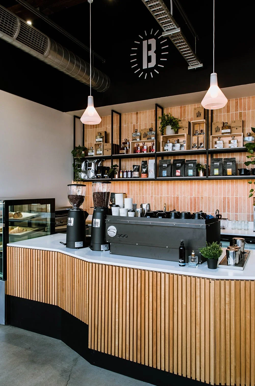 Interior view of a modern coffee shop with a wooden counter, coffee grinders, and shelves stocked with coffee bags and jars, hanging pendant lights, and a large clock on the ceiling.
