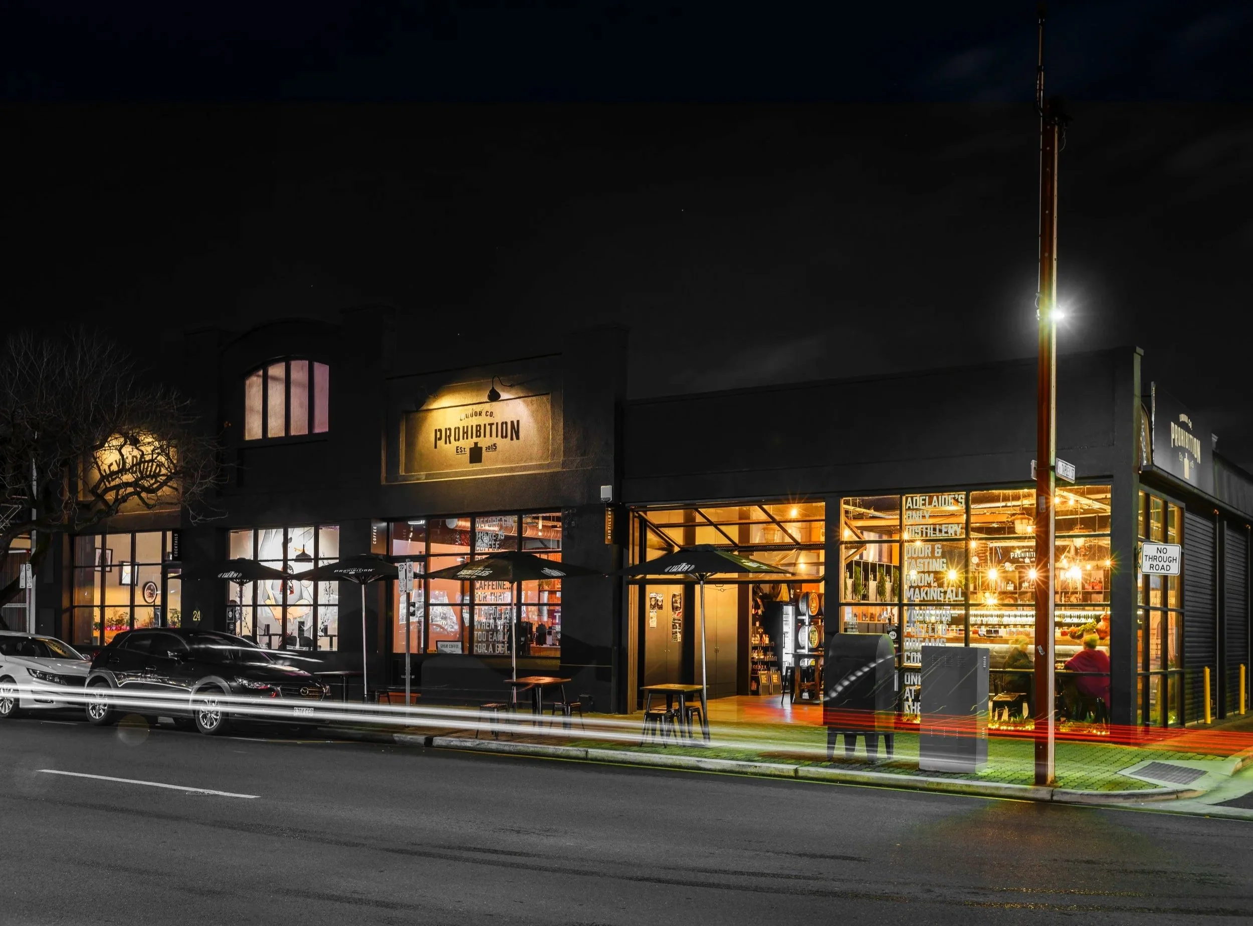 Nighttime street view of a storefront with large glass windows and a sign that reads 'Prohibition'. The interior is illuminated, revealing shelves and a bar area. In front, there is a parking lot with a few parked cars, and some outdoor seating with umbrellas. A tall streetlight is visible on the right side of the image.