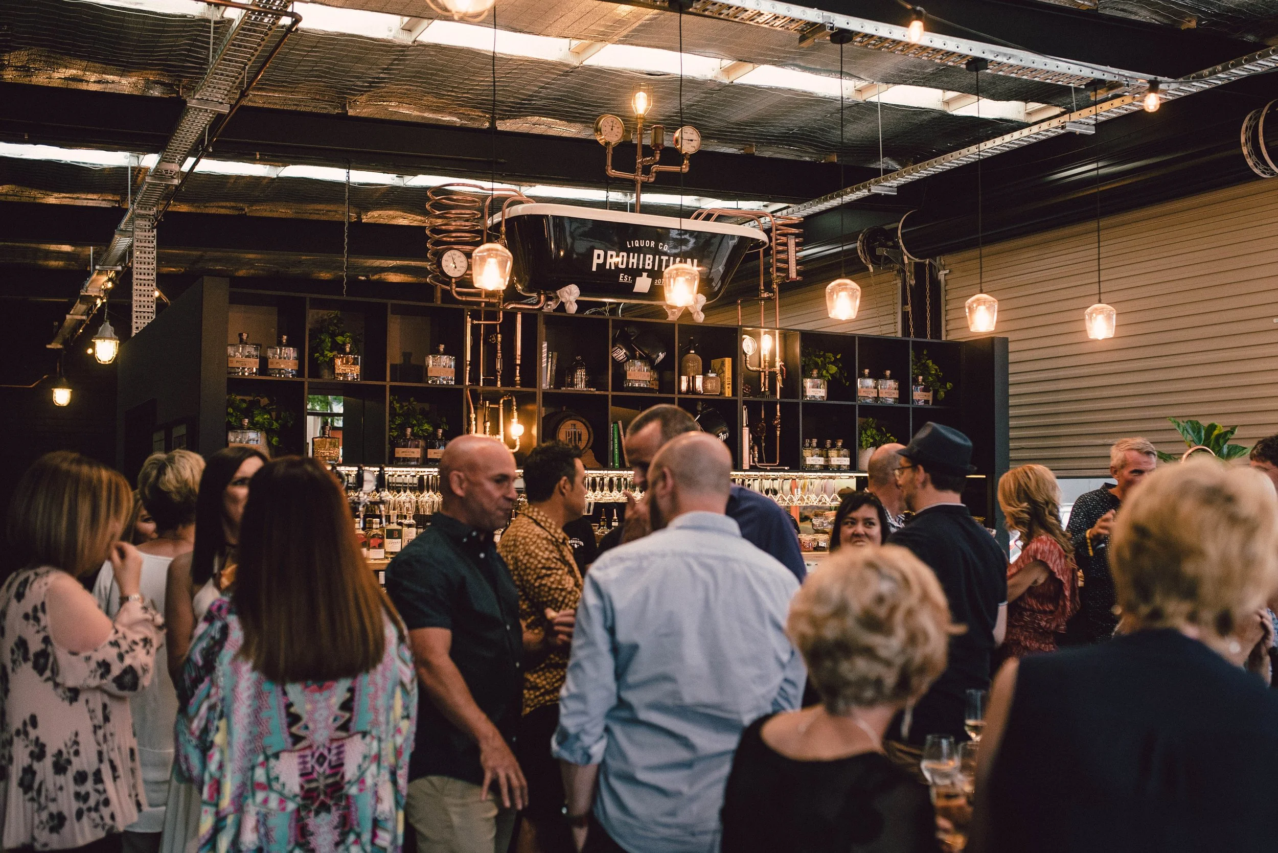 A crowded indoor bar or restaurant with people socializing. The ceiling has industrial decor with exposed pipes and hanging lights. A large black sign with white text reads 'LIQUOR CO PROHIBITION EST. 201' mounted on the ceiling. Shelves behind the bar display bottles and plants.