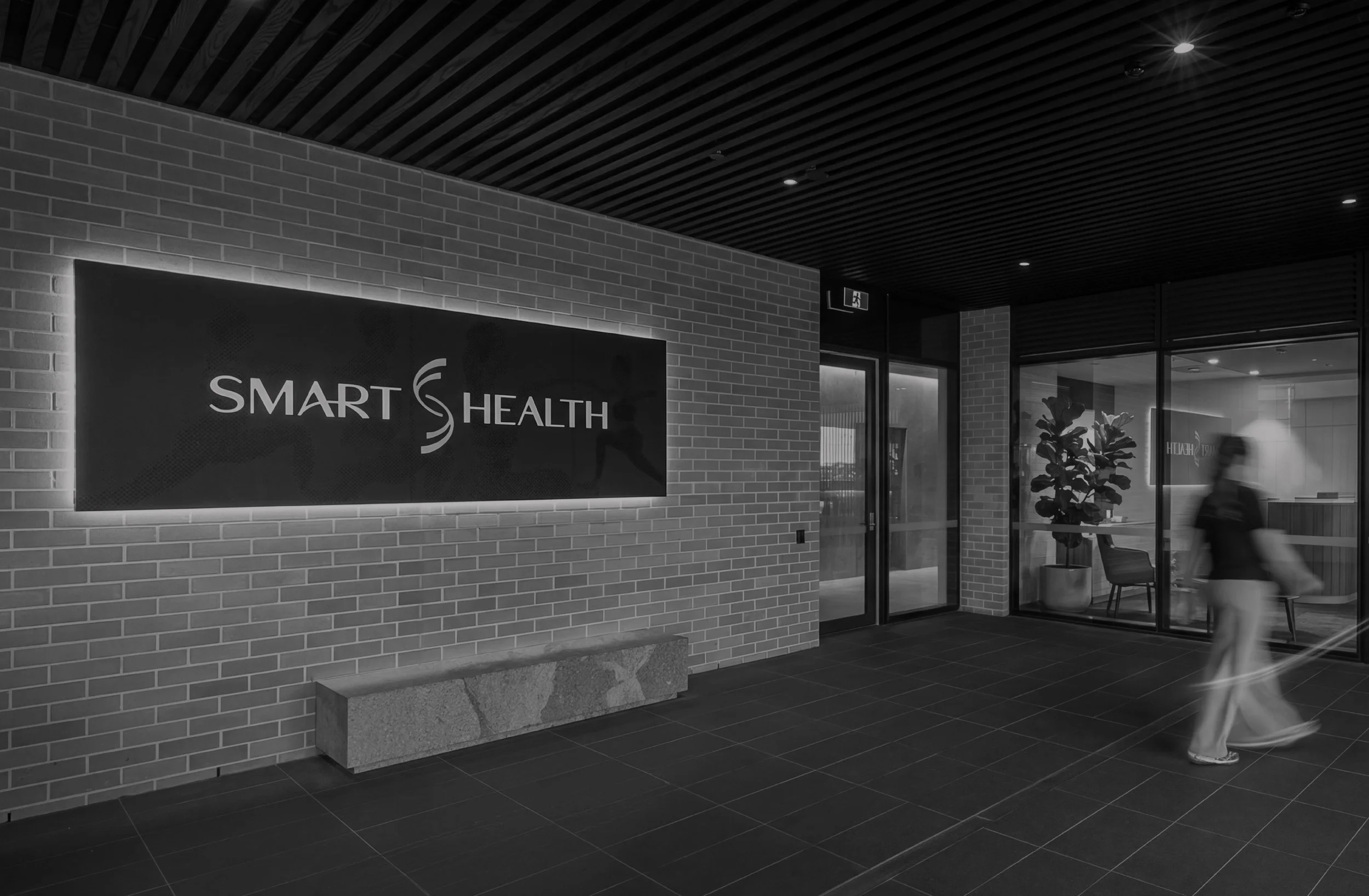 Interior view of a modern healthcare building lobby with a brick wall, a sign that reads 'Smart Health', glass doors, a person walking, potted plant, ceiling with recessed lighting.