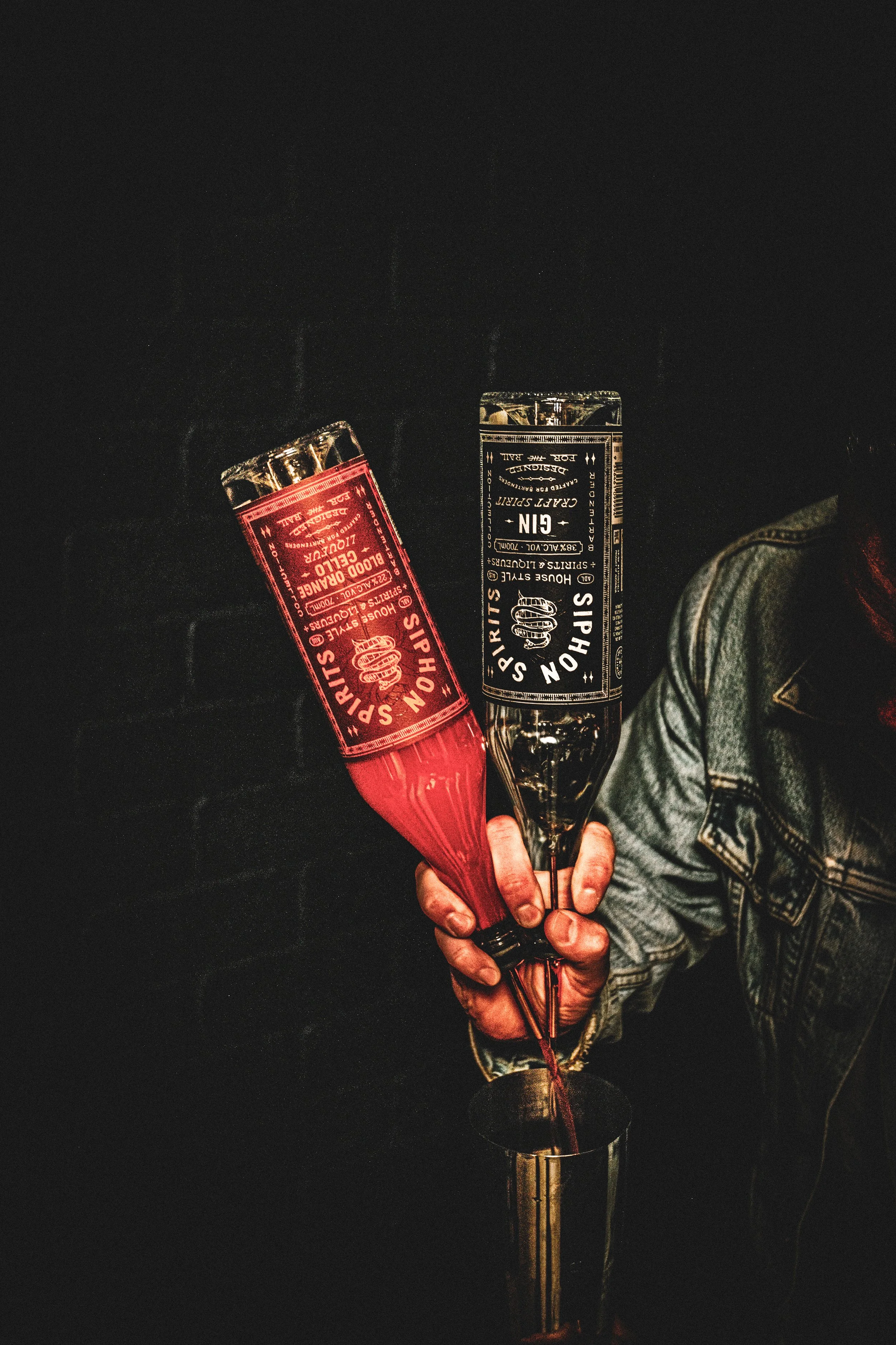 Two people holding cocktails, one in a red glass and one in a clear glass, against a dark background. One person is wearing a denim jacket.