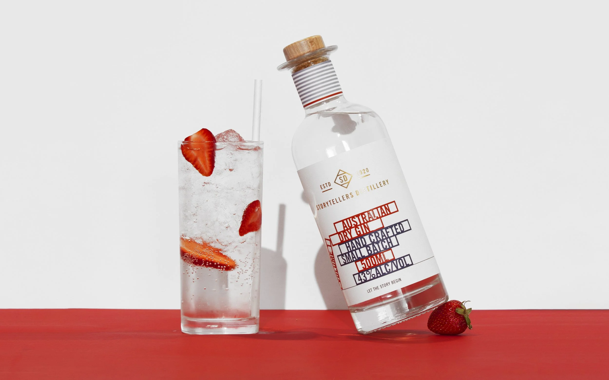 A glass of sparkling water with strawberries and ice, next to a bottle of Australian dry gin on a red surface against a white background.