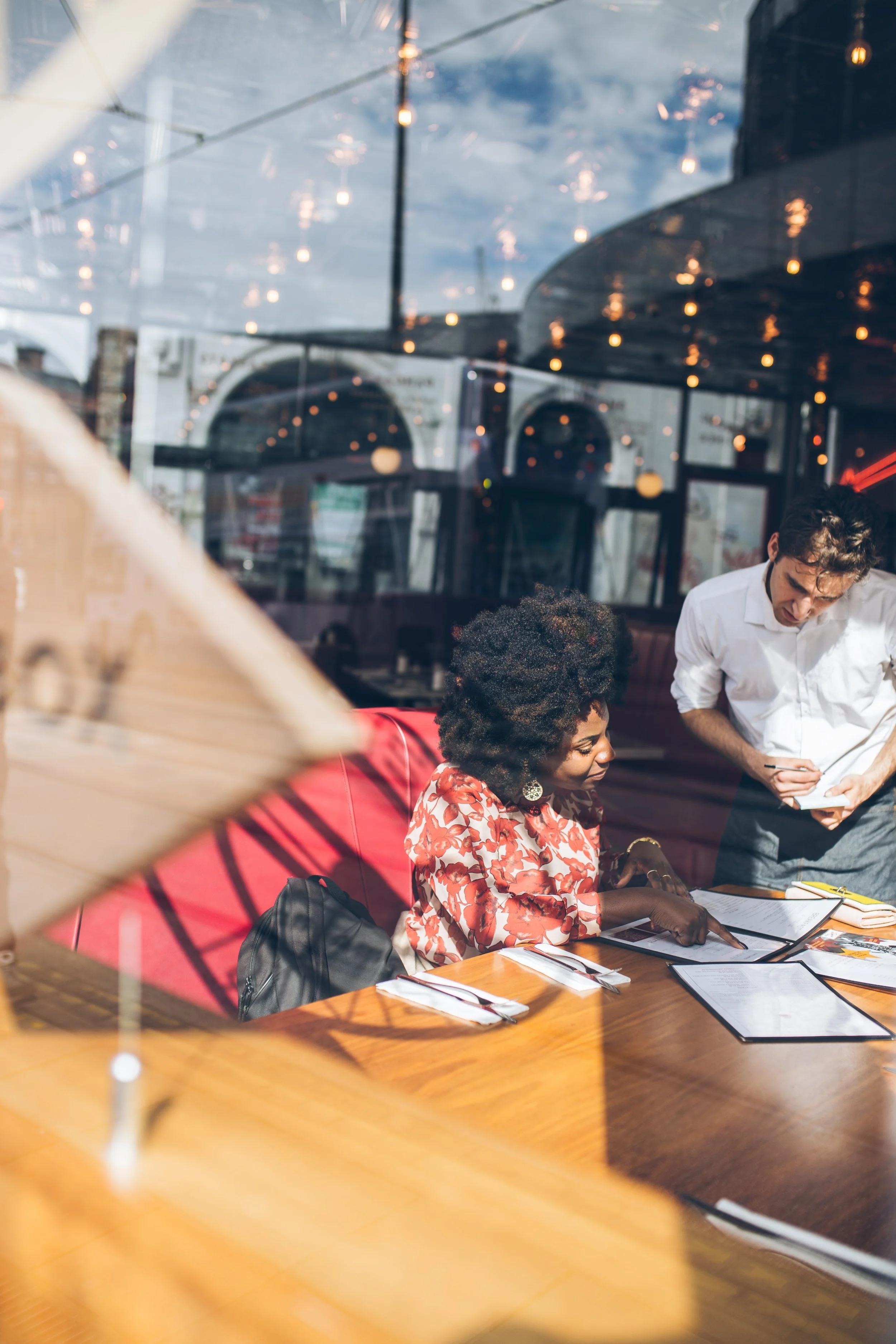 Two people in a restaurant, looking at menus on the table while talking to each other, viewed through a glass window.