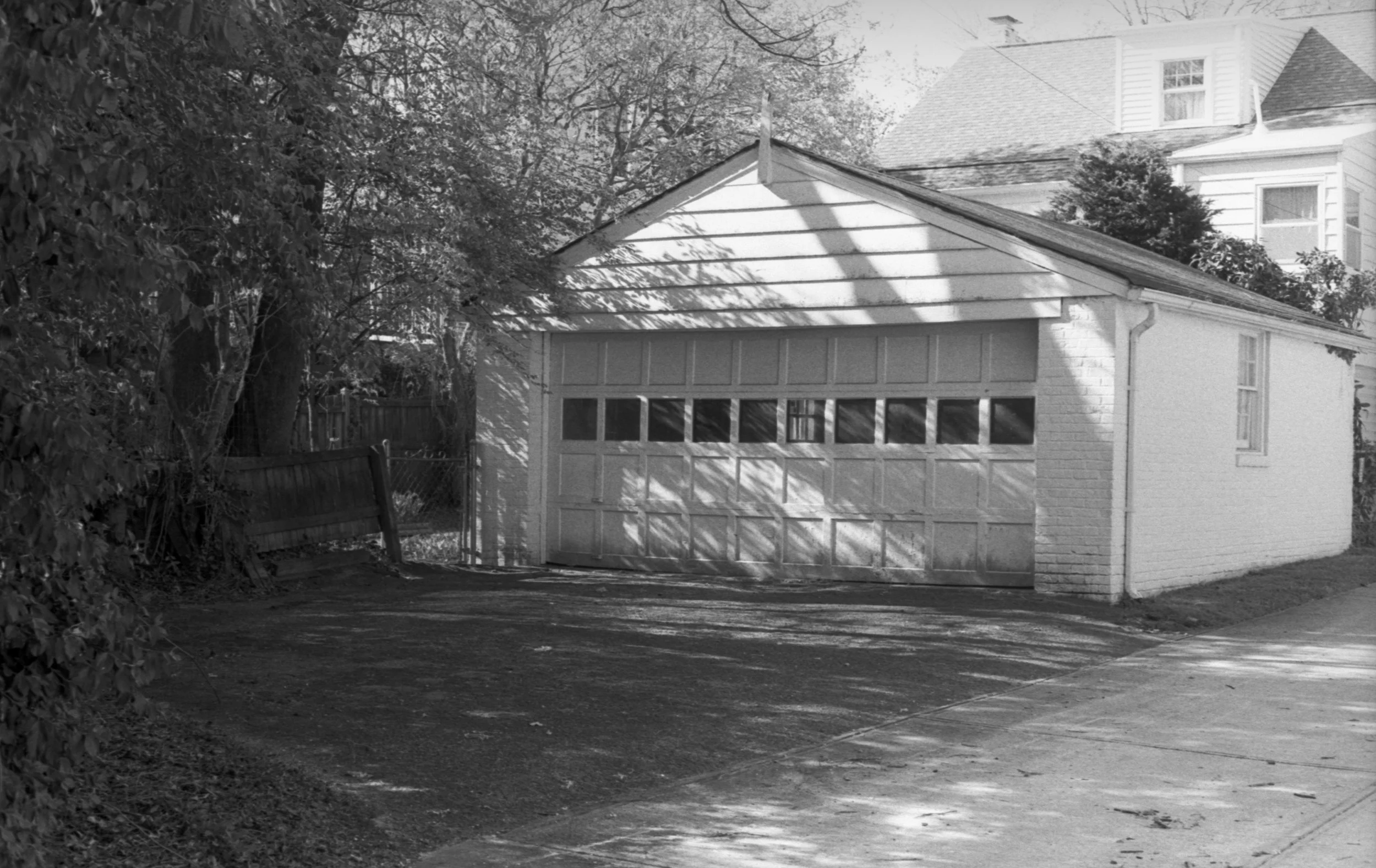 Black and white photo of a small garage attached to a house, with a large tree on the left casting shadows on the garage roof and yard, and a wooden fence on the left side.