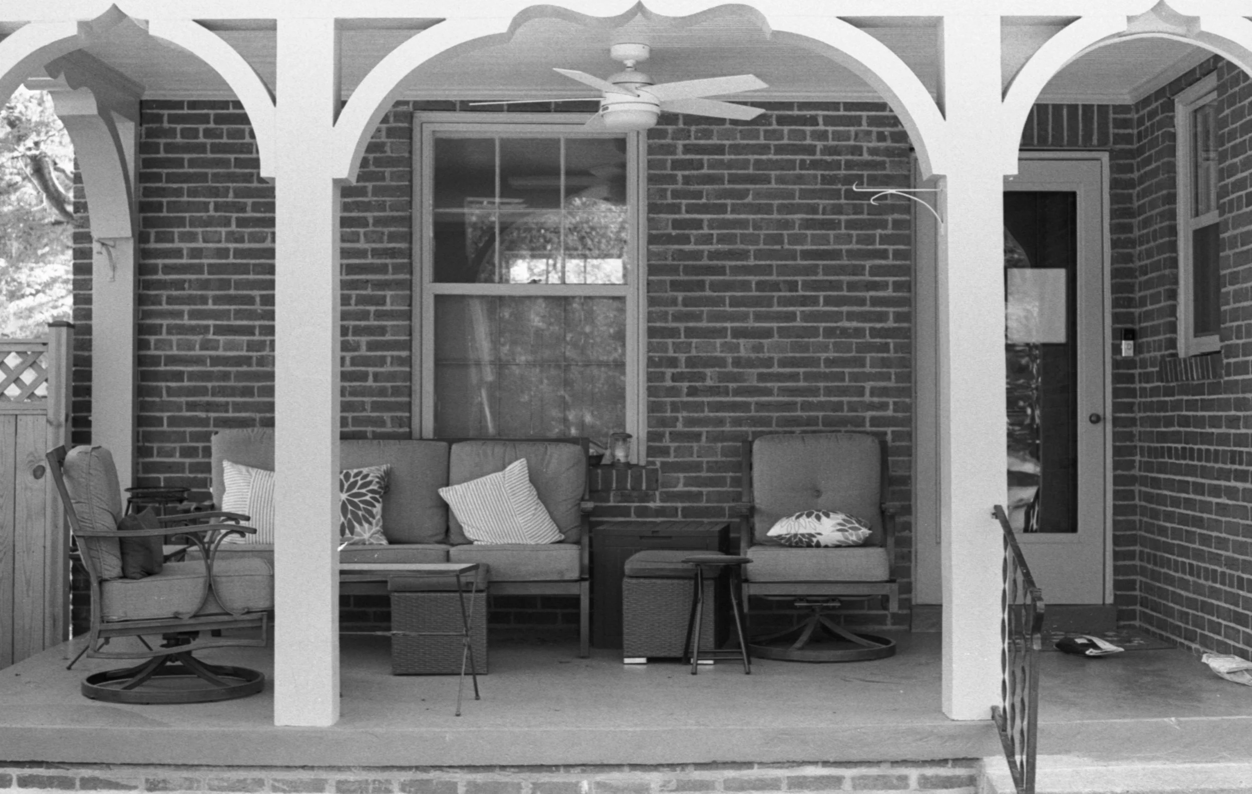 Black-and-white photo of a covered porch with brick walls and white arches, furnished with outdoor seating including cushioned chairs, pillows, a small table, and a ceiling fan.