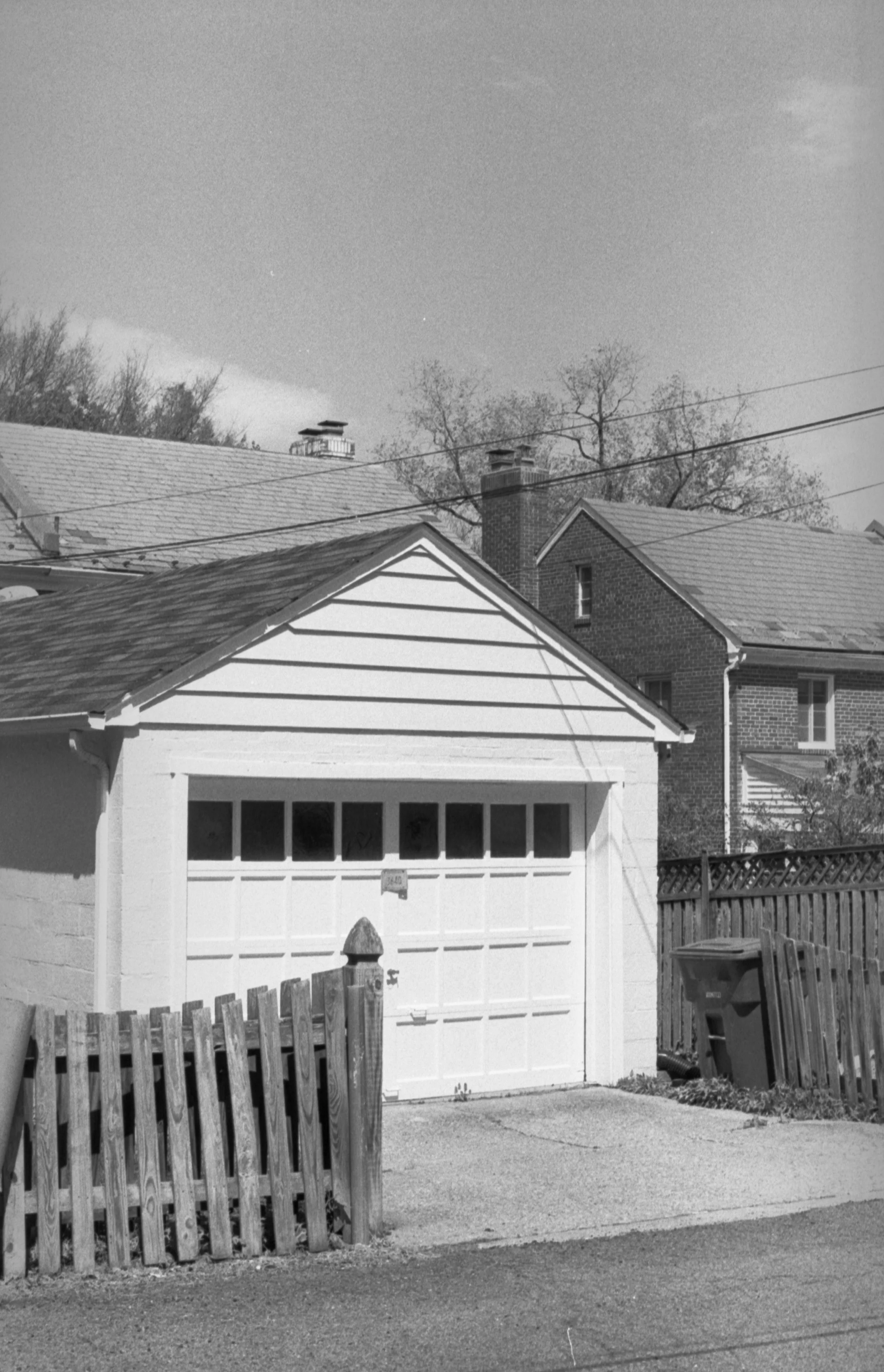 Black and white photo of a suburban house with a garage, a wooden fence, and trees in the background under a clear sky.