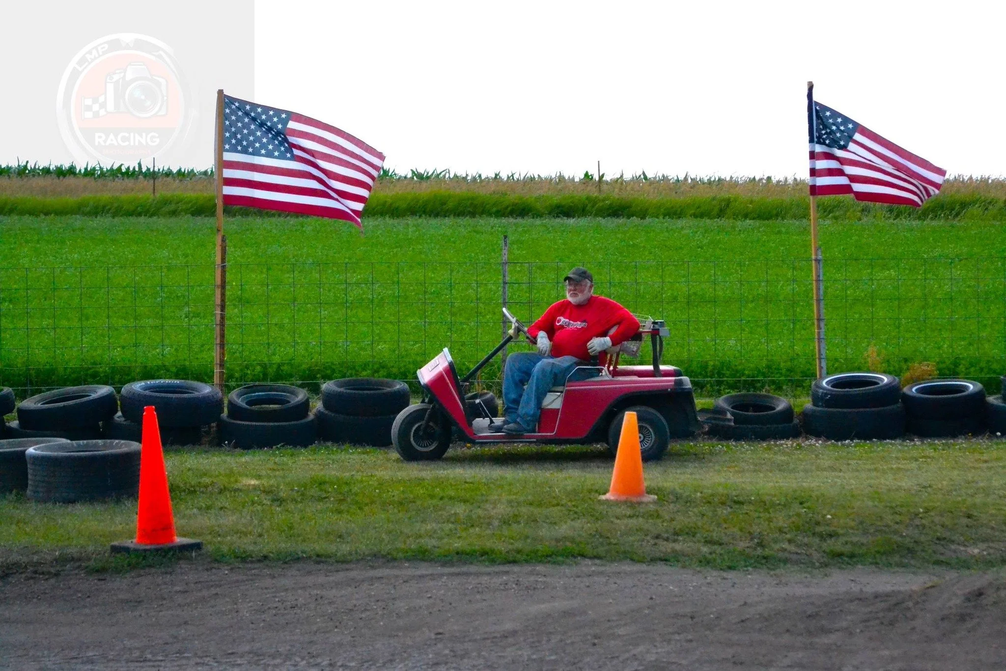 A man in a red shirt and jeans sitting in a red golf cart near a fence with two American flags, orange cones, and black tires, with a green field and blue sky in the background.