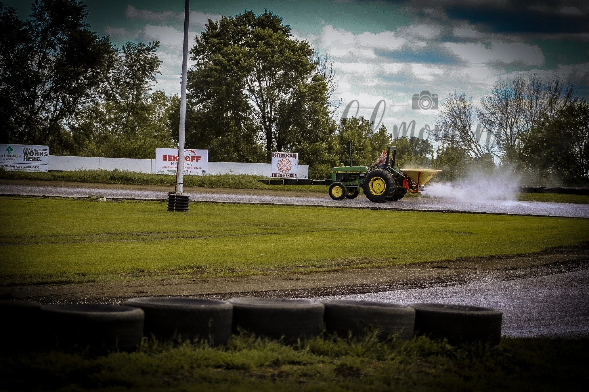 A tractor is seen on a race track with spray behind it, surrounded by trees and with advertisements on a fence in the background.