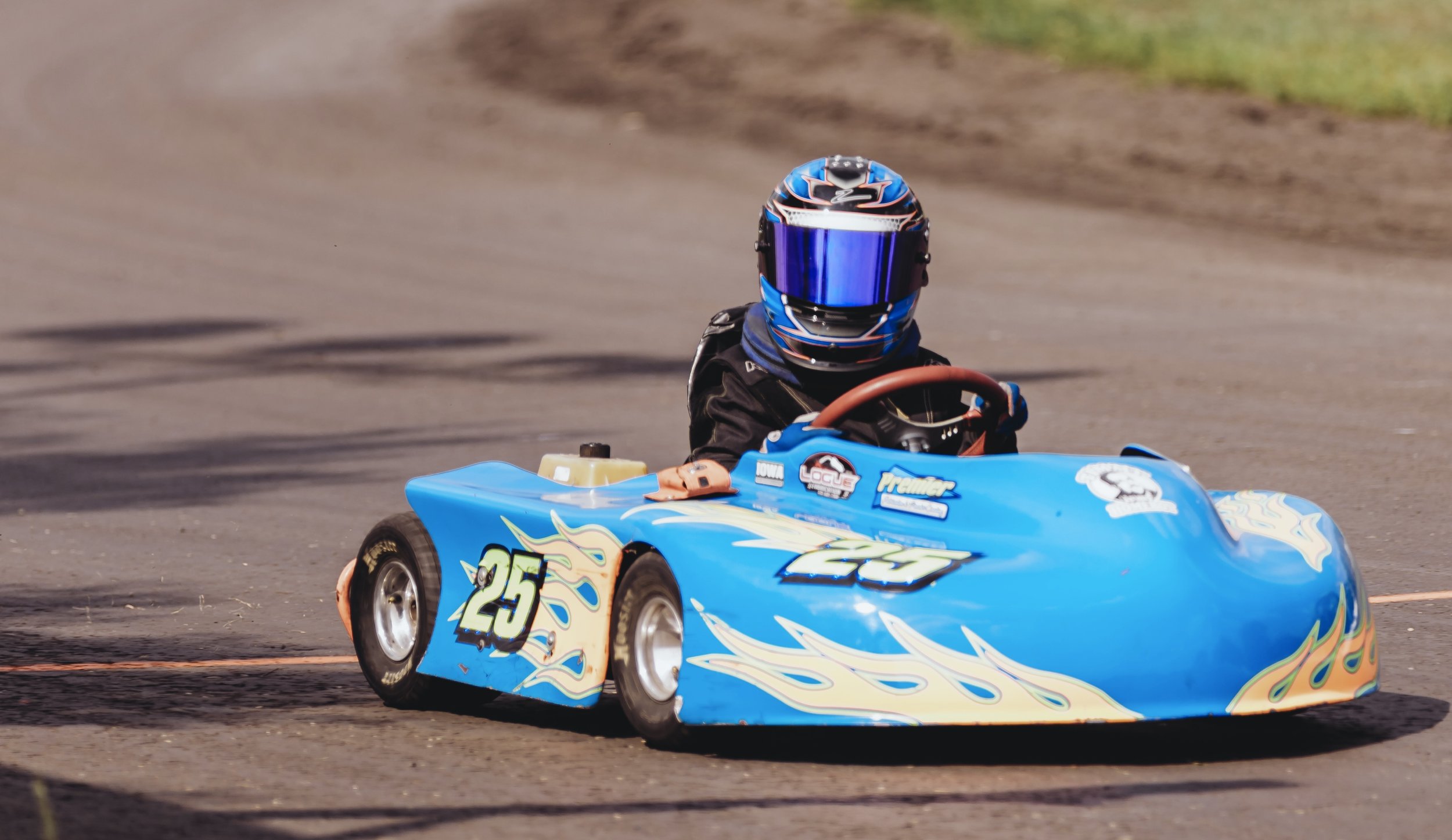 A go-kart racer wearing a blue helmet and black racing suit drives a blue go-kart on a dirt track. The go-kart has the number 25 with flames decals on the sides.