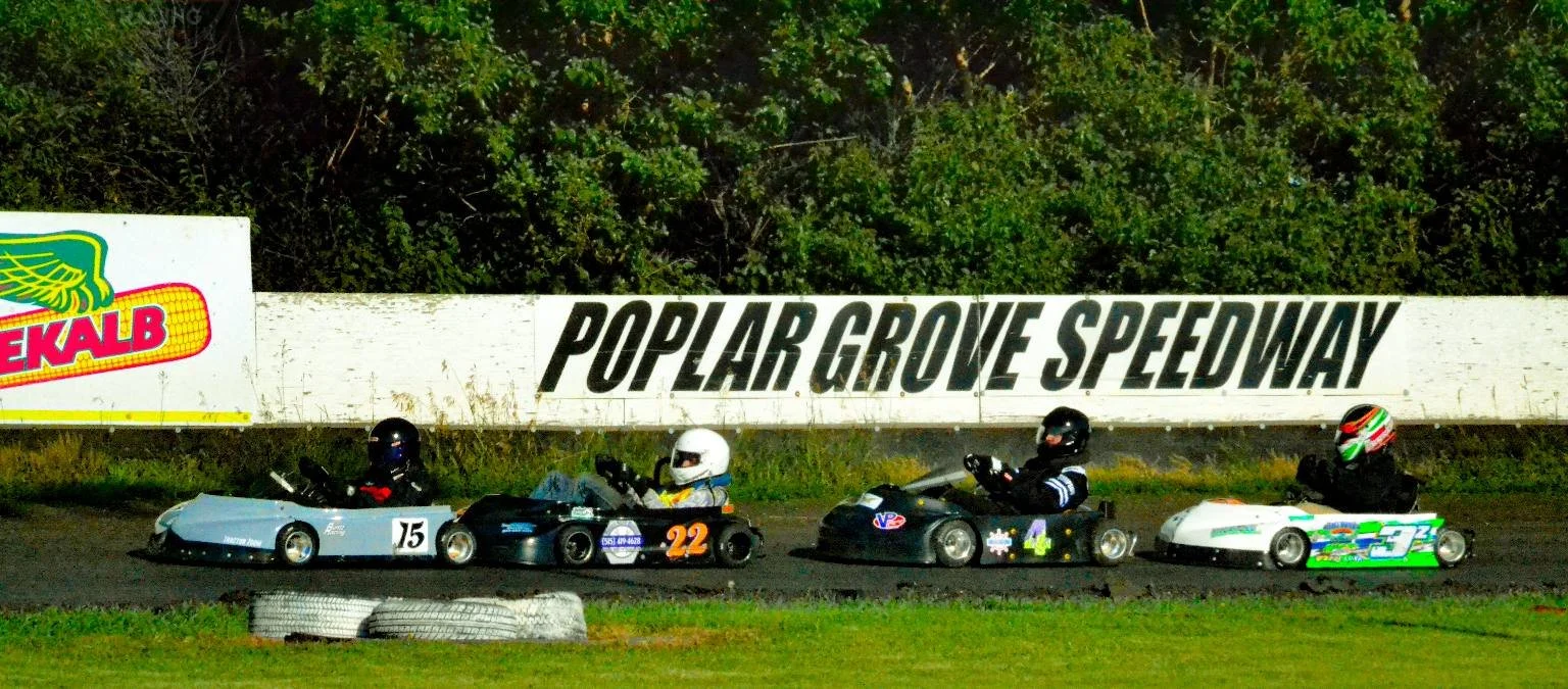 Four go-karts racing on a track at Poplar Grove Speedway, with drivers wearing helmets and racing suits, and a sign for the speedway in the background.
