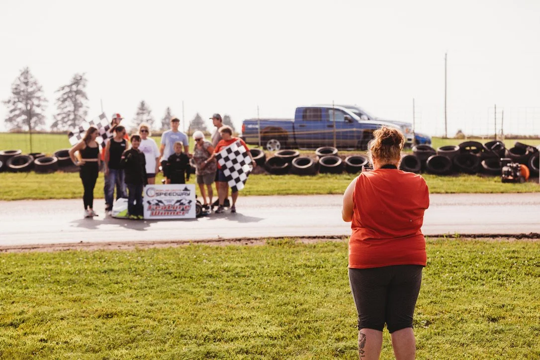 A group of people at a race track, some holding checkered flags and a banner, with a woman in a red shirt taking a photo in the foreground.